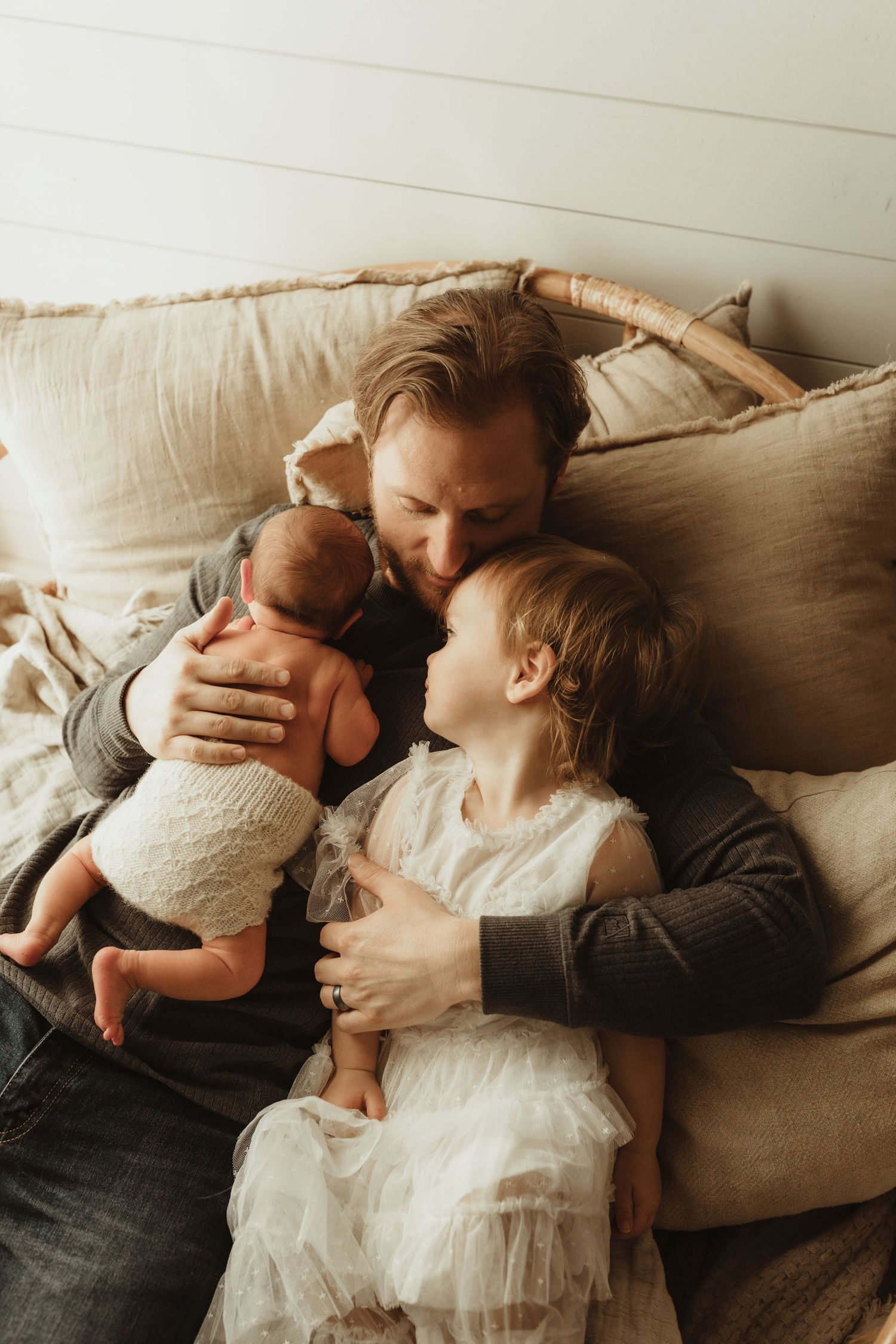 Father cuddles newborn baby and young daughter in white dress on cozy beige cushioned bed.
