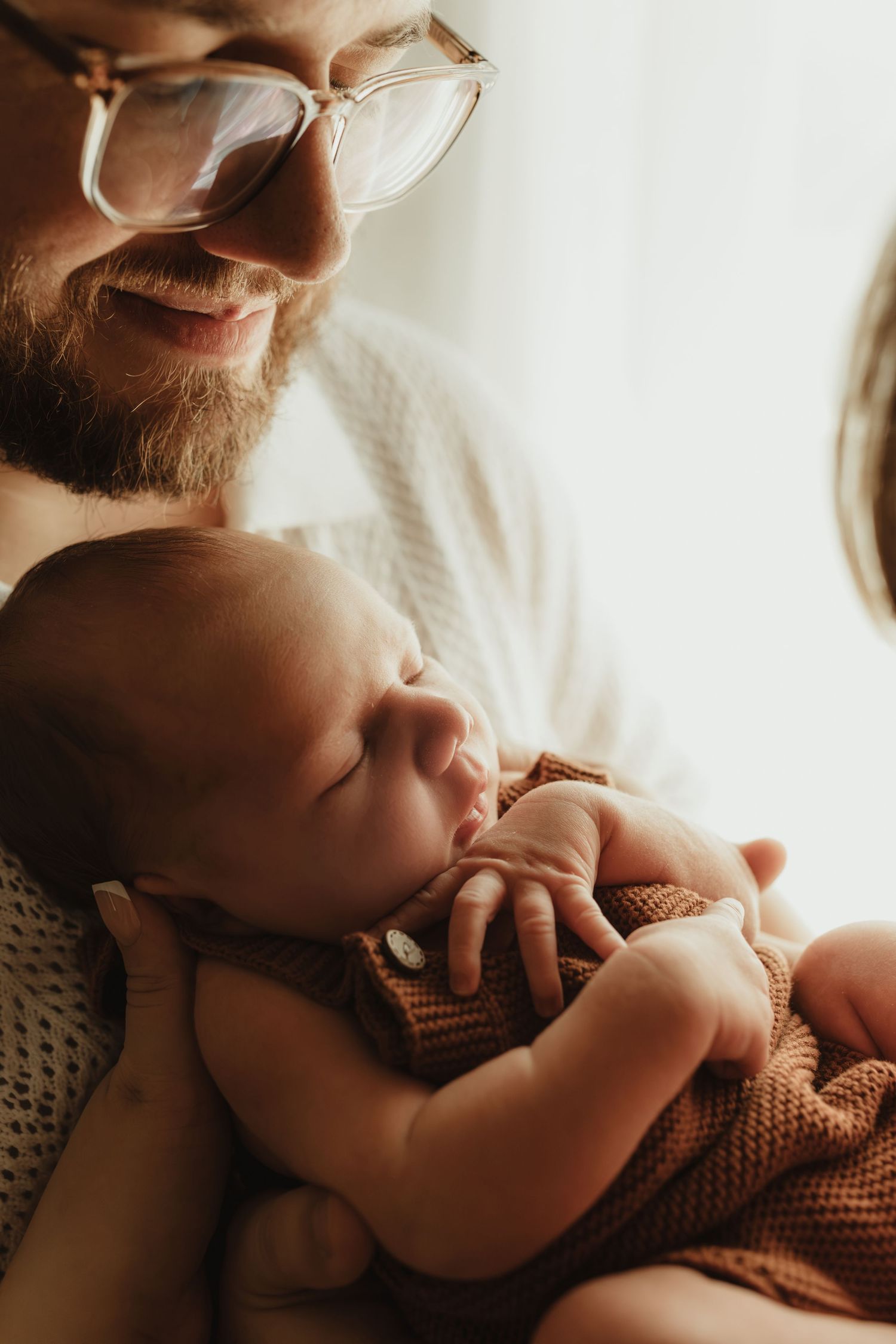 Smiling bearded man with glasses tenderly holding a sleeping newborn baby dressed in a rust-colored knit outfit.