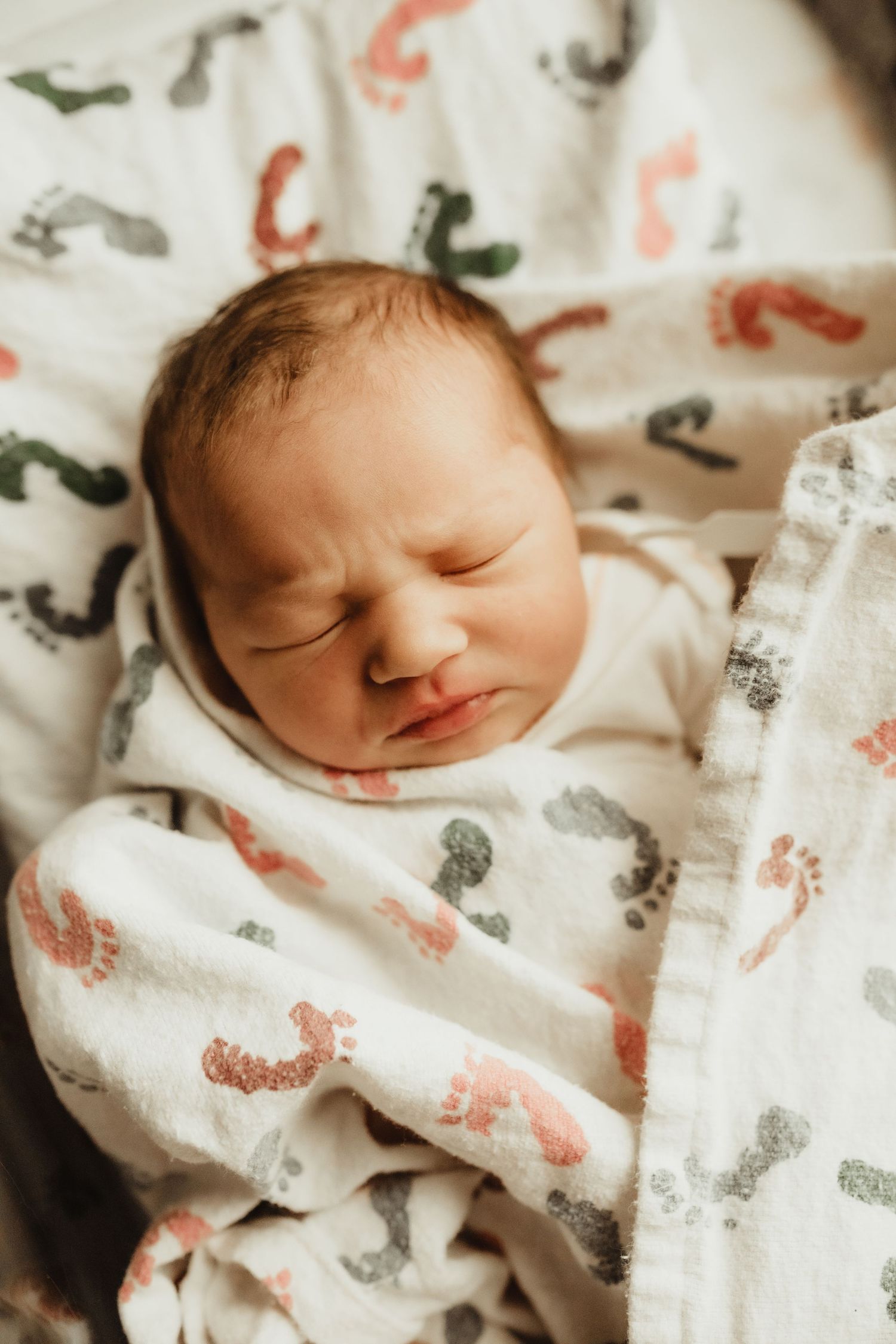 Peaceful newborn baby sleeping swaddled in a white muslin blanket with colorful footprint patterns.