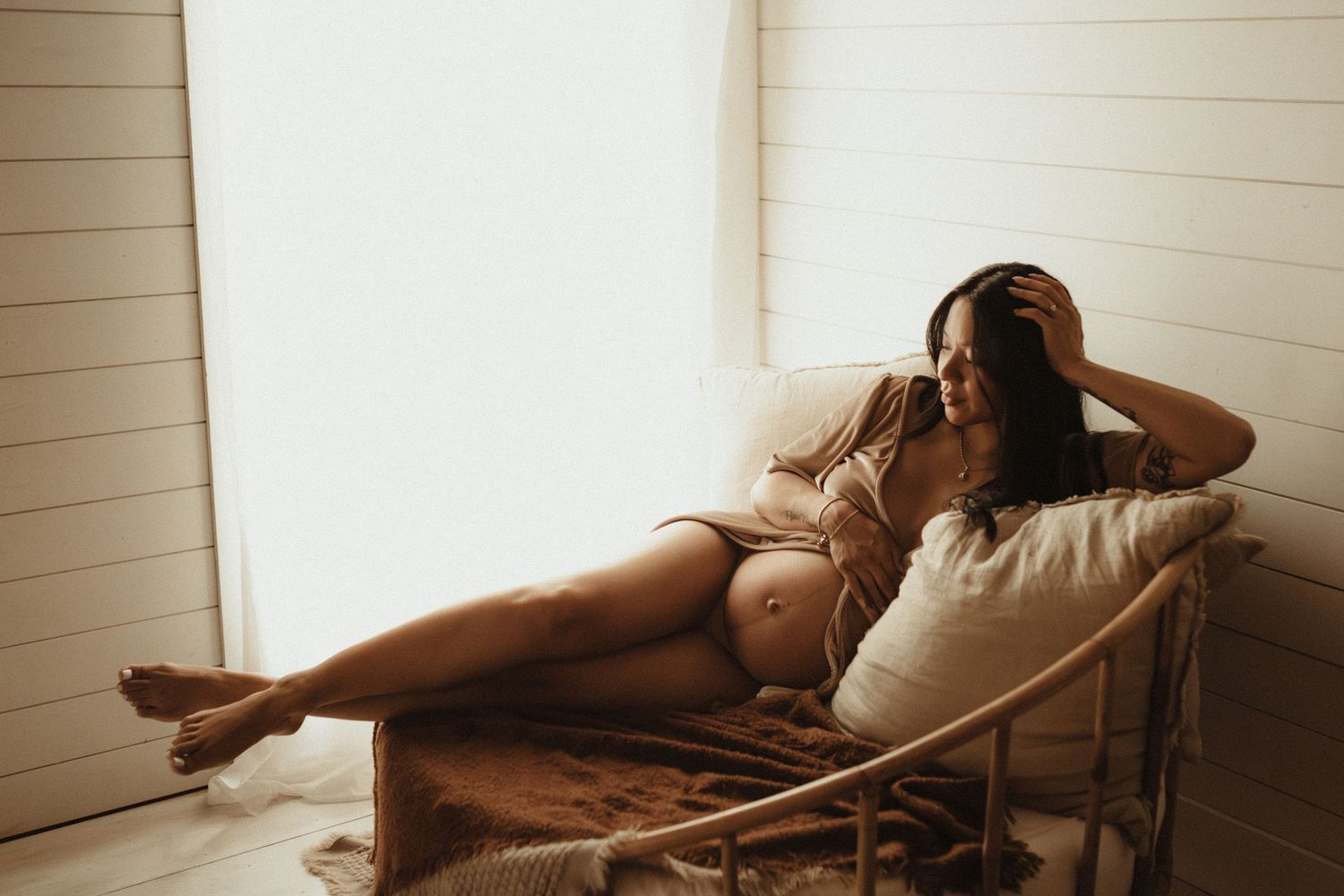 Pregnant woman in beige lingerie reclines on a chair by a bright window, smiling and cradling her baby bump.
