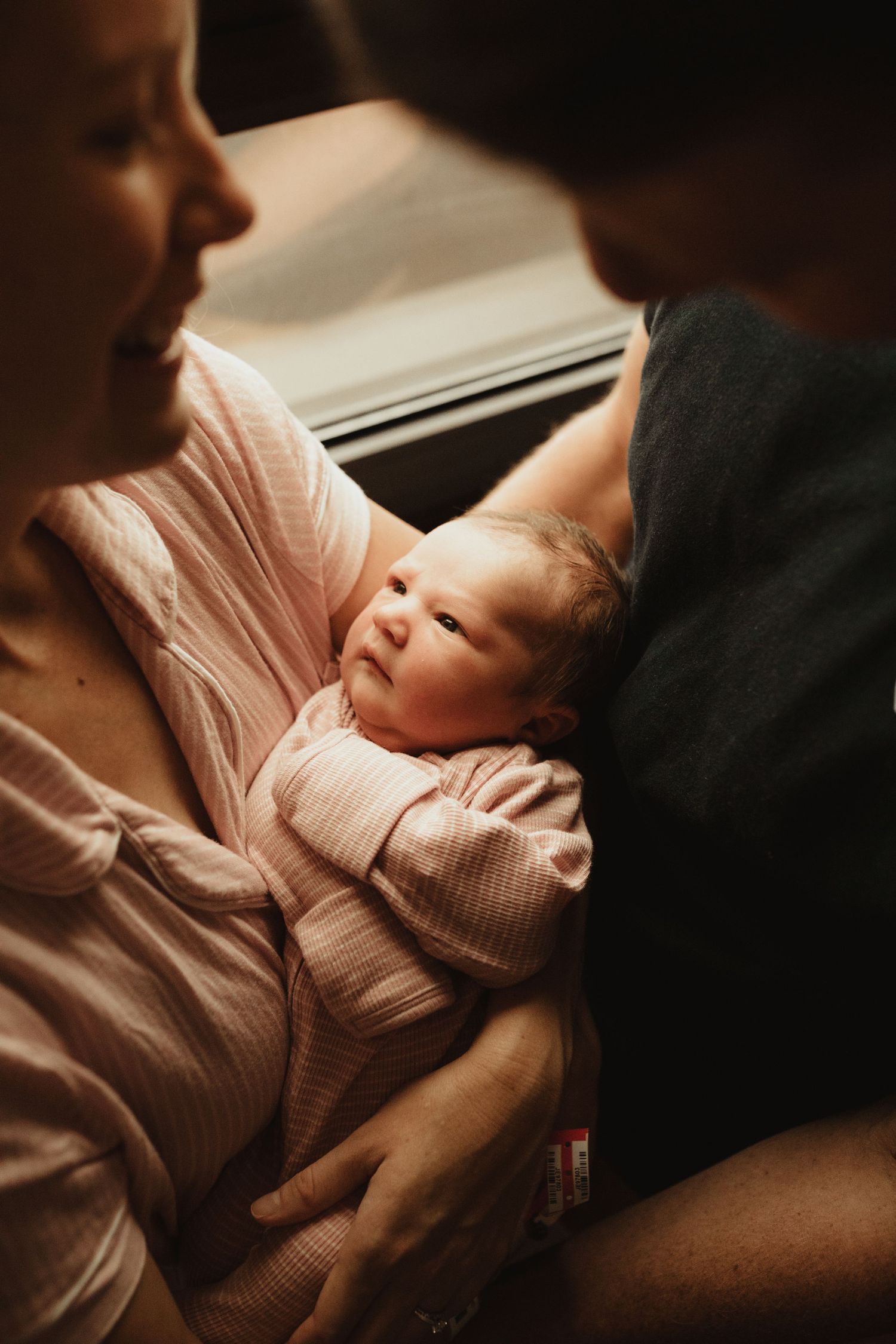Mother tenderly holds her newborn baby dressed in pink against her chest near a window.