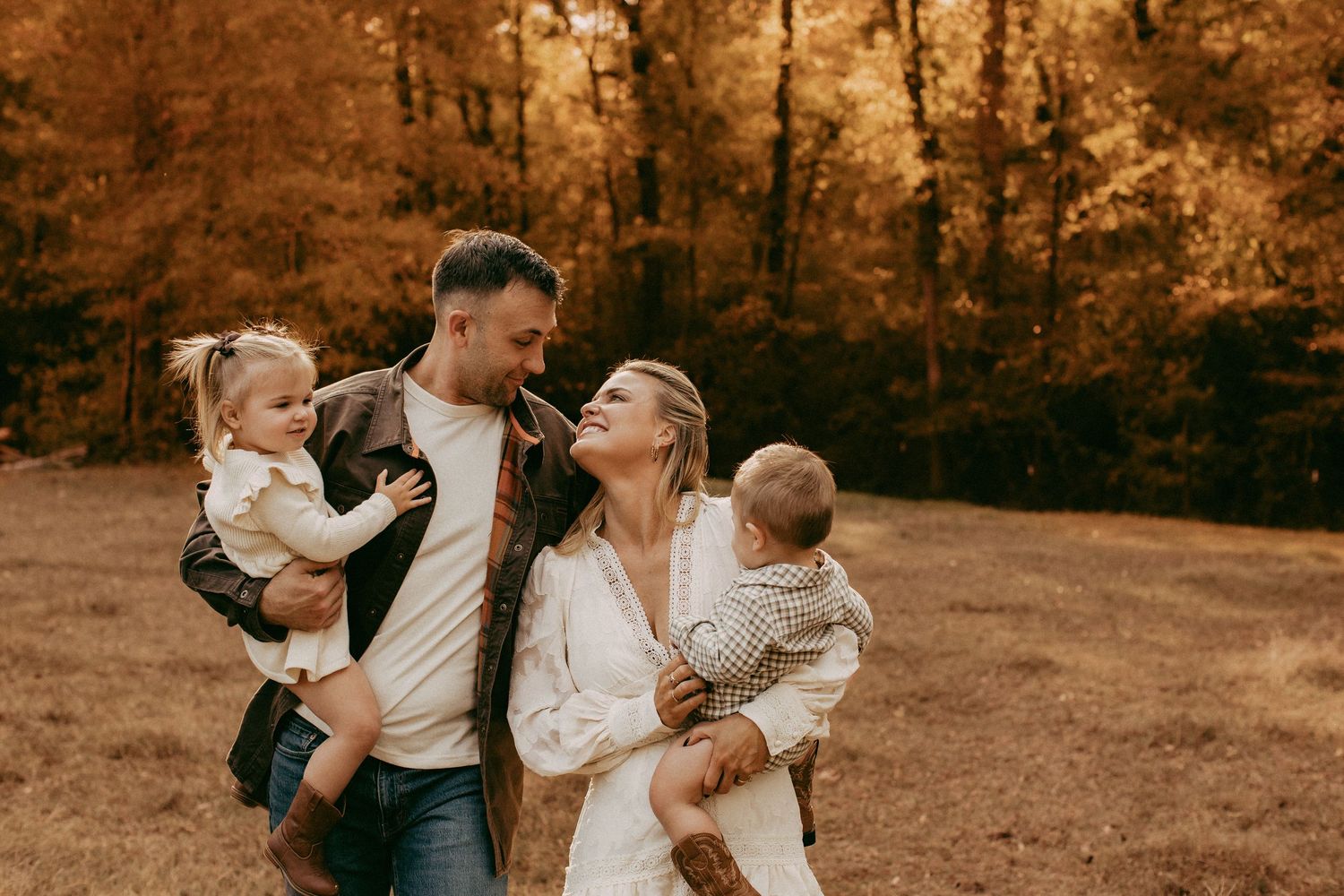 Family of four enjoying an autumn outdoor photoshoot, parents holding toddlers among golden fall foliage.