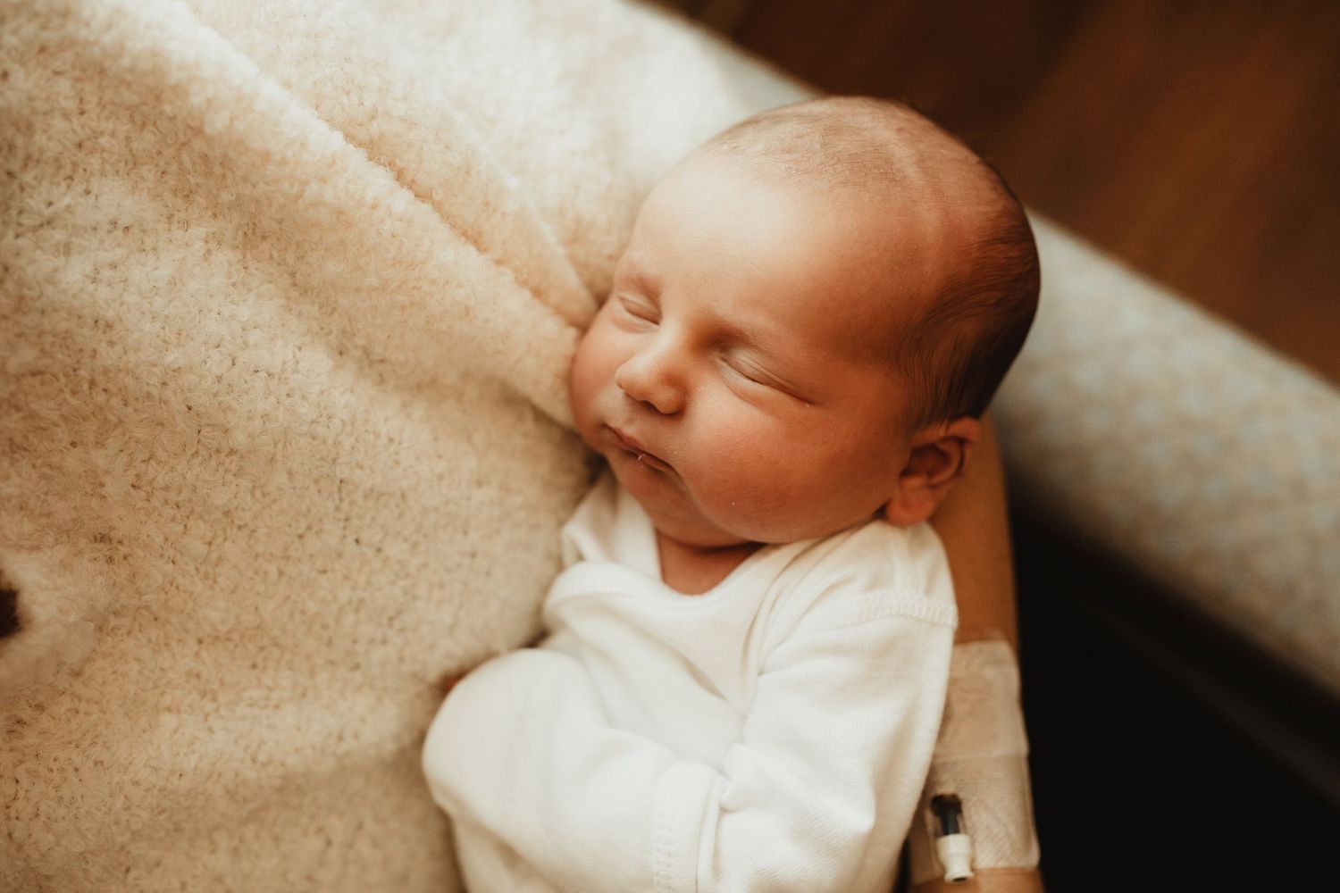 A small infant wearing white sleepwear rests peacefully on cream-colored bedding during naptime.