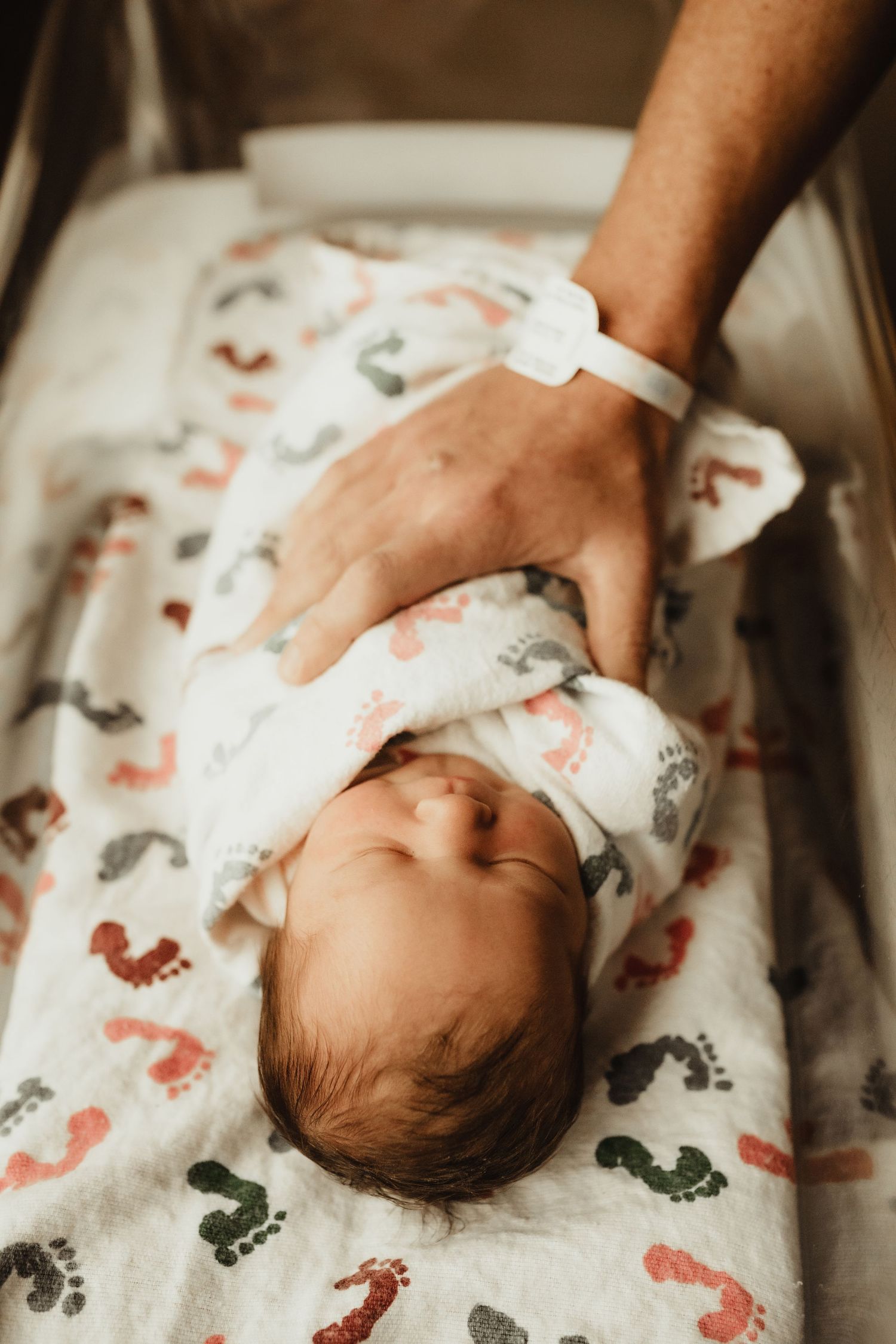 Newborn baby swaddled in a footprint blanket in a hospital bassinet with an adult hand gently resting on them.