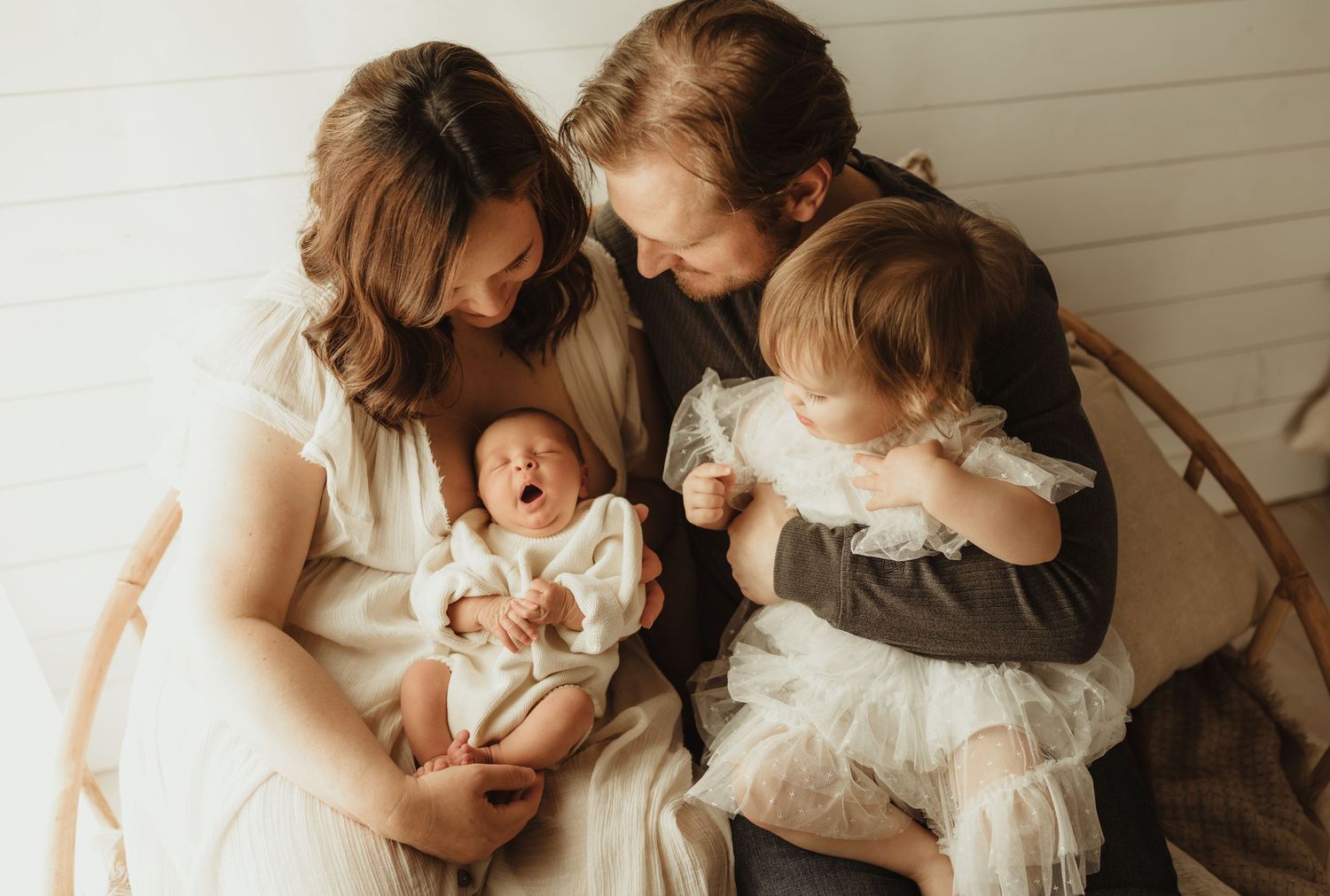 A young family of four sits together, parents cradling a newborn baby while a toddler in a white dress looks on.