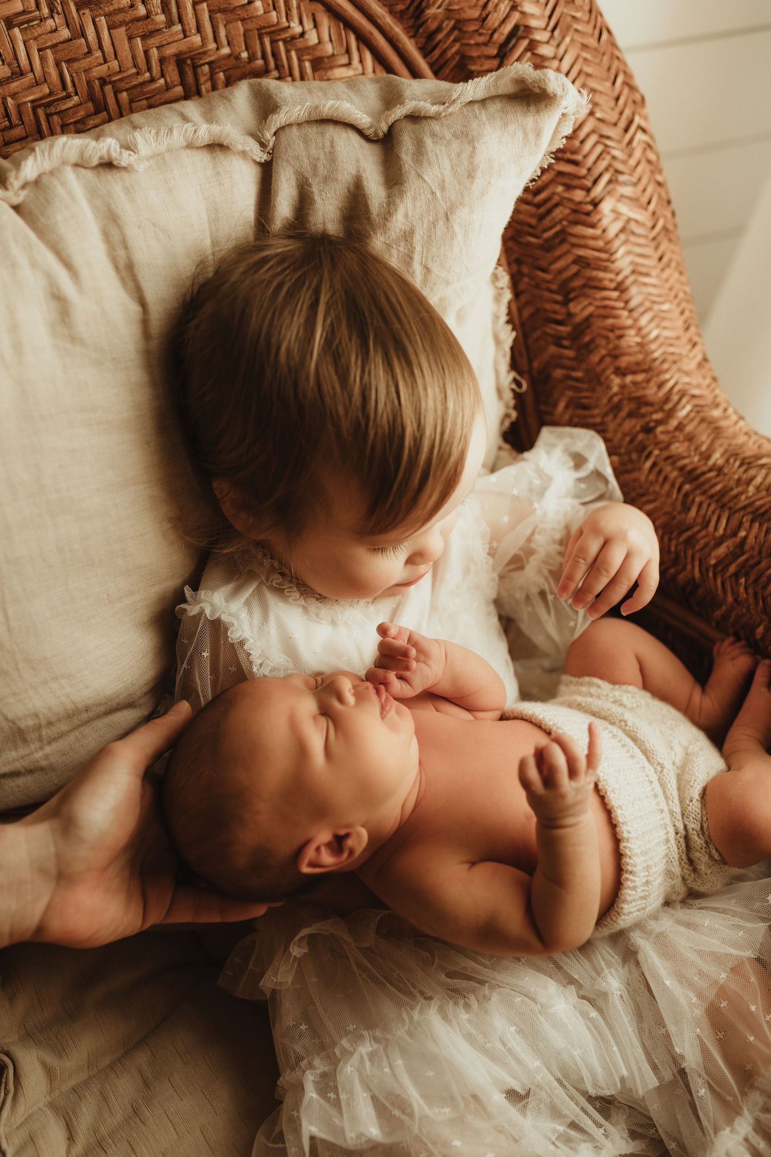 A toddler in a white dress gently kisses a sleeping newborn baby in a wicker chair with soft linen cushions.