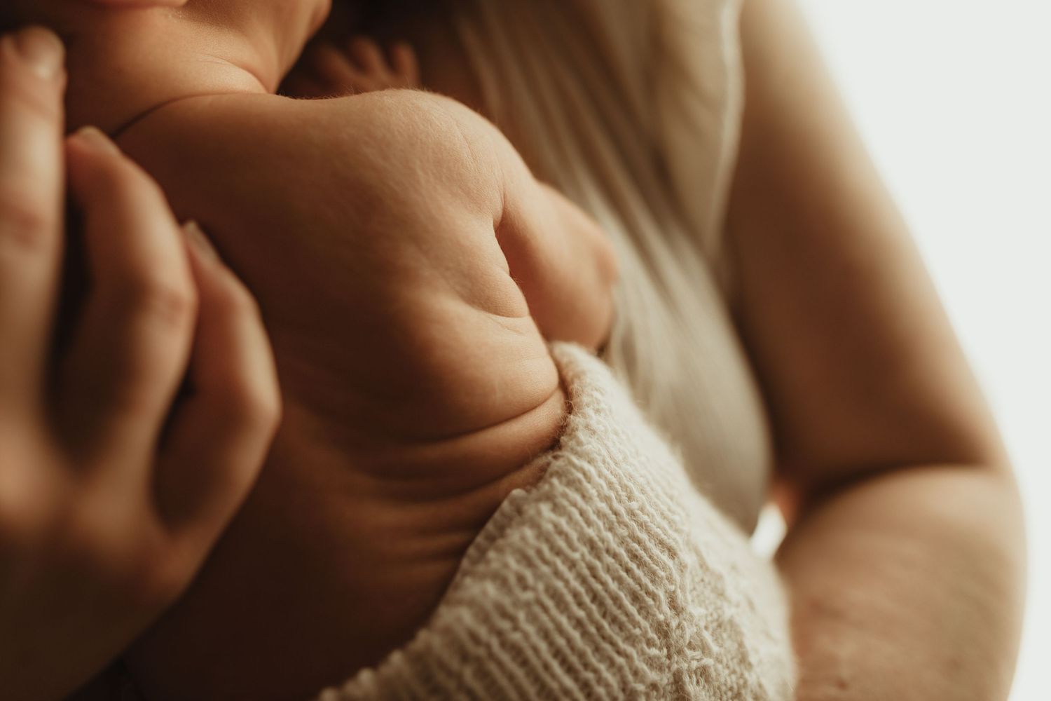 Close-up of a newborn baby's tiny hand resting gently on a parent's arm in soft, warm lighting.