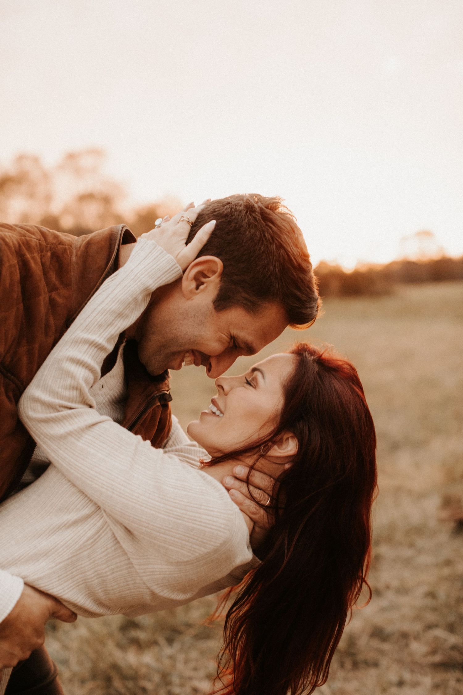 Couple sharing a romantic dip kiss in a golden field at sunset, woman with red hair smiling up at man.