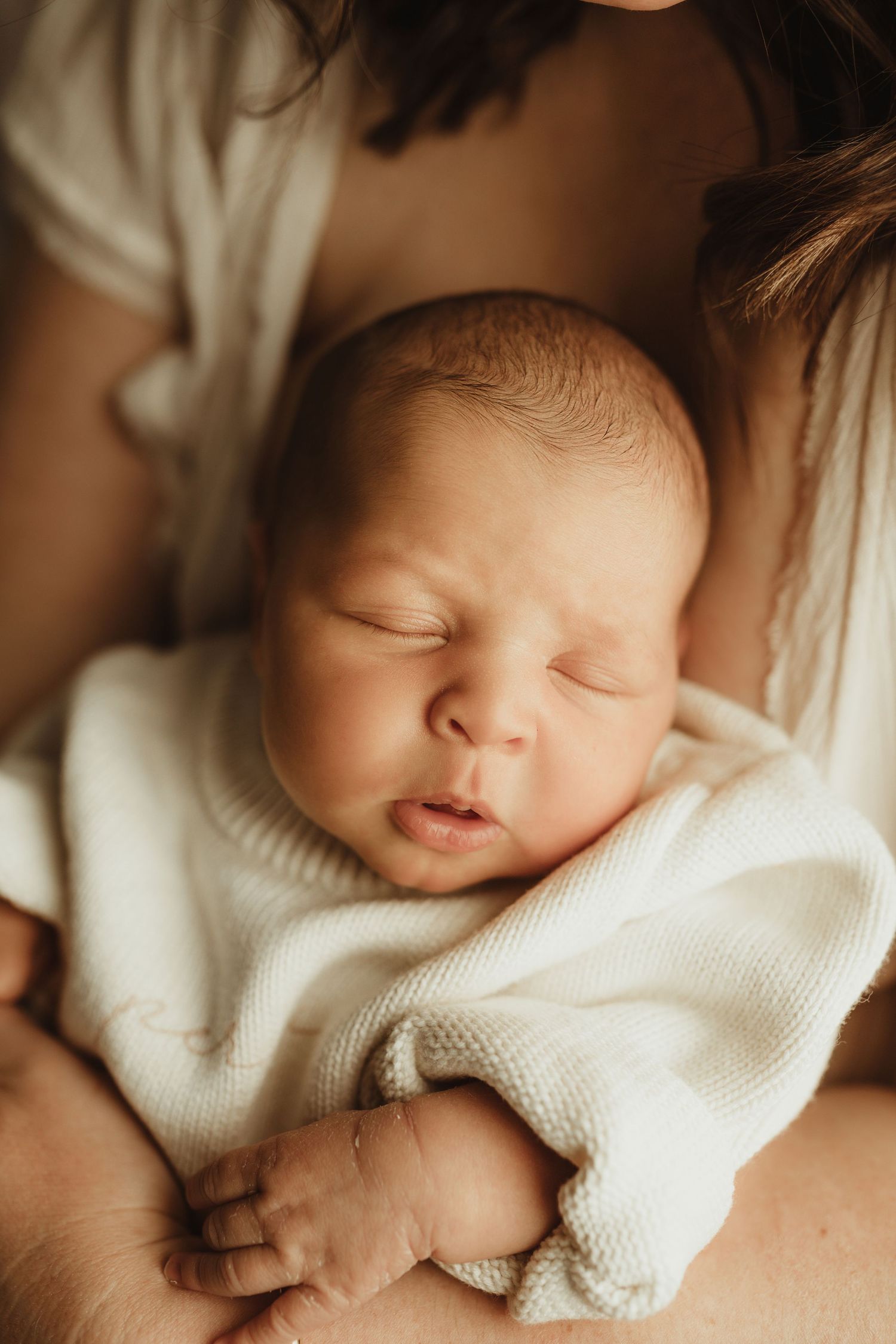 A peaceful newborn baby sleeping in a mother's arms, wrapped in a soft white knit blanket.