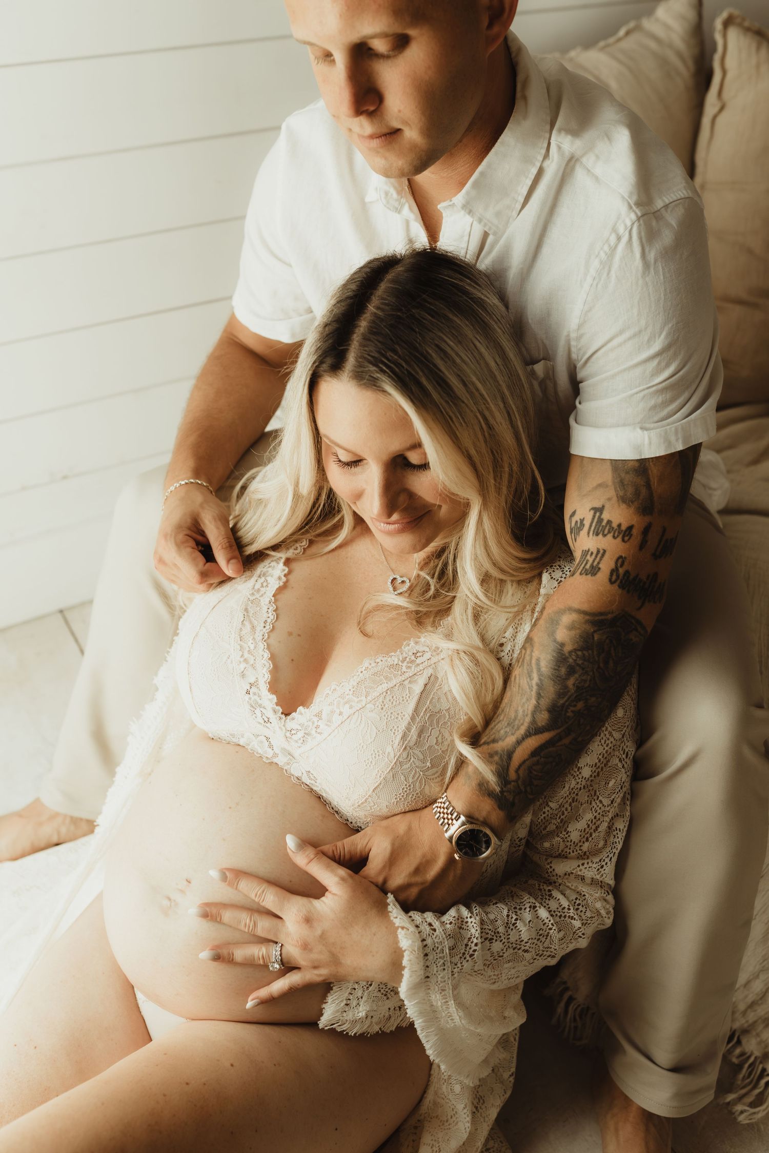 Couple in white maternity photo shoot, pregnant woman in lace robe cradled by tattooed partner on bed.