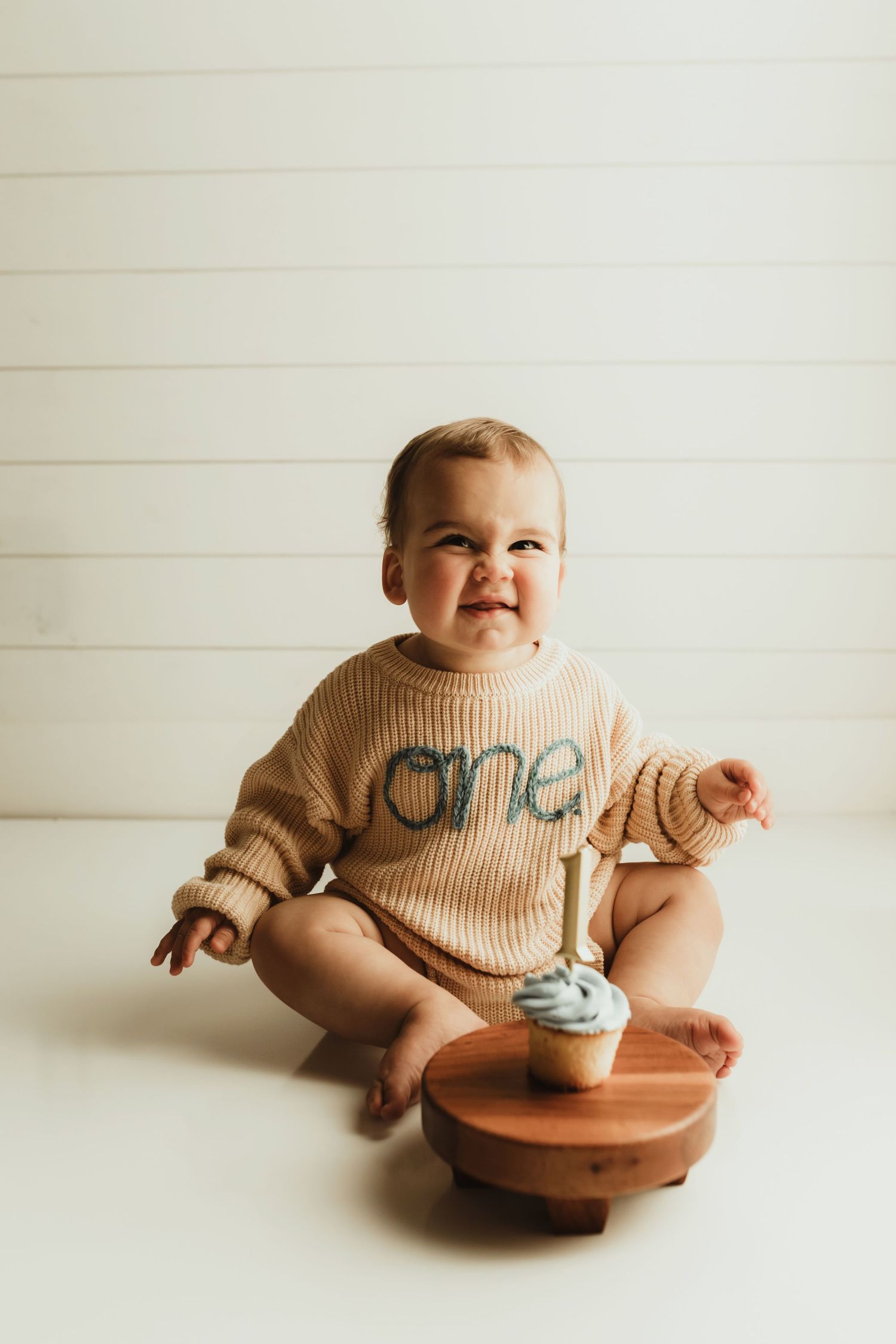 A baby wearing a beige knit sweater sits and smiles while playing on a wooden stool against a white background.