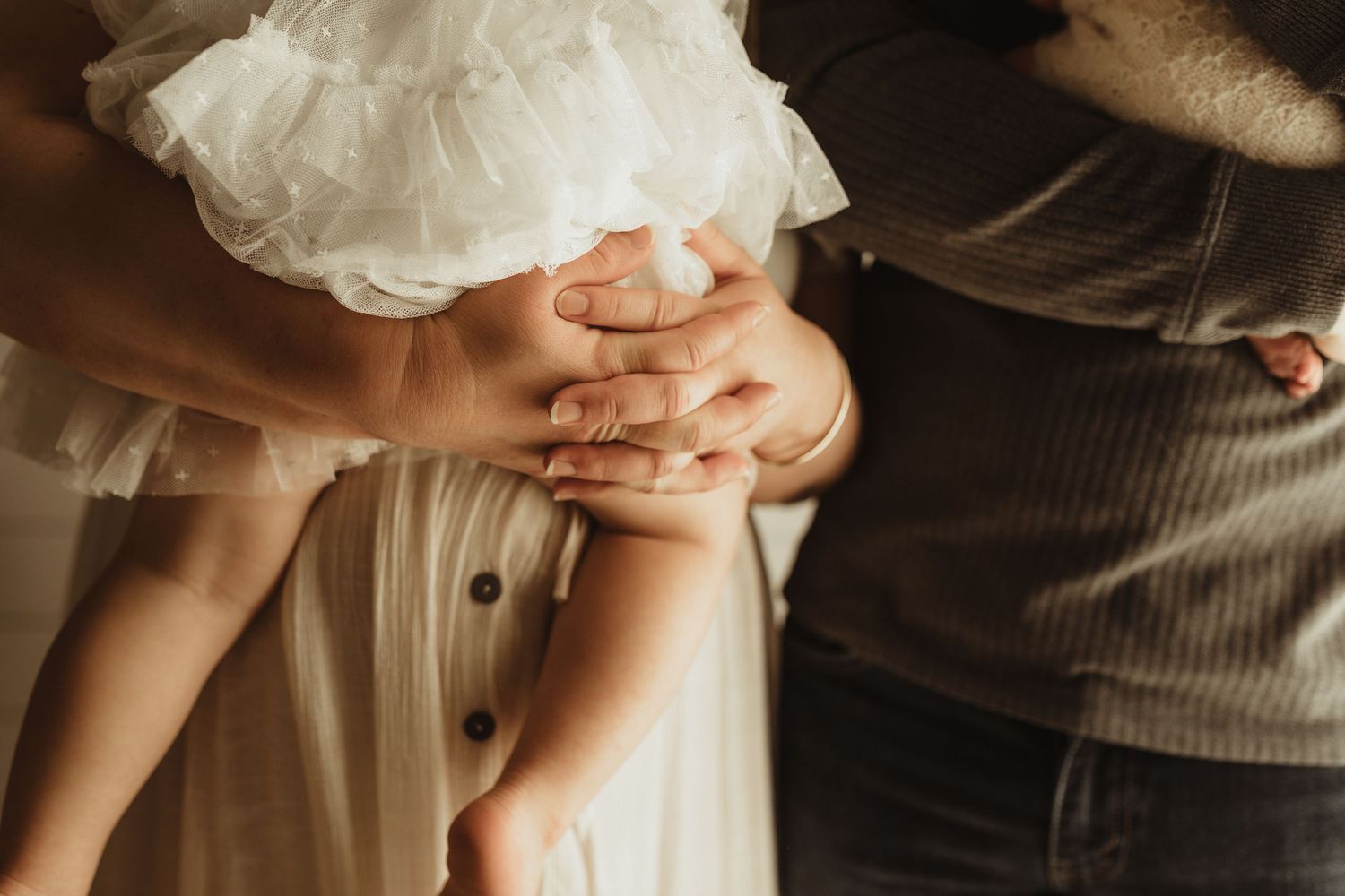 Parents holding their baby's hands in a tender family moment, with a child in a white dress.