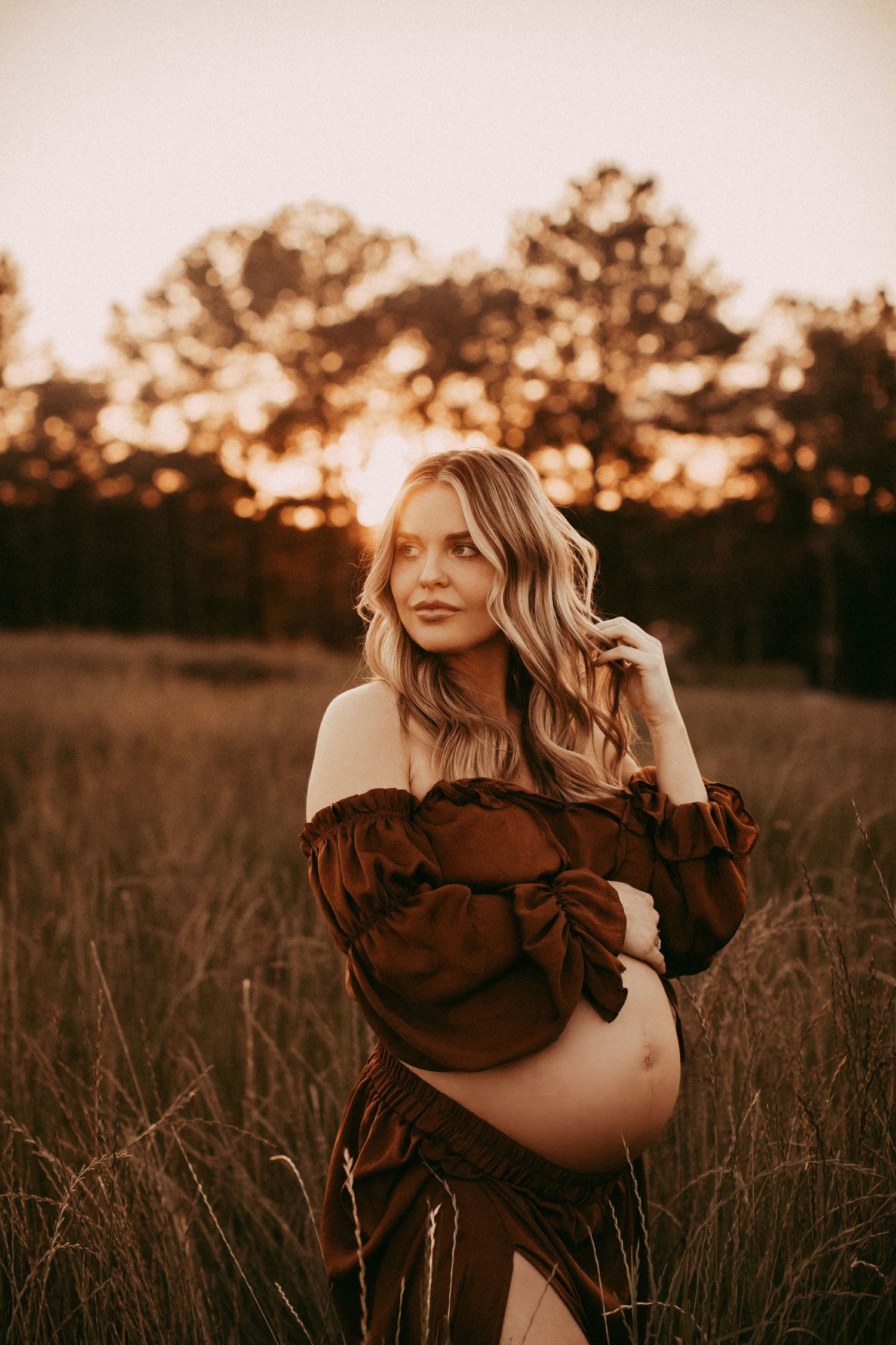A person in a brown off-shoulder dress poses in a golden field at sunset with flowing hair.