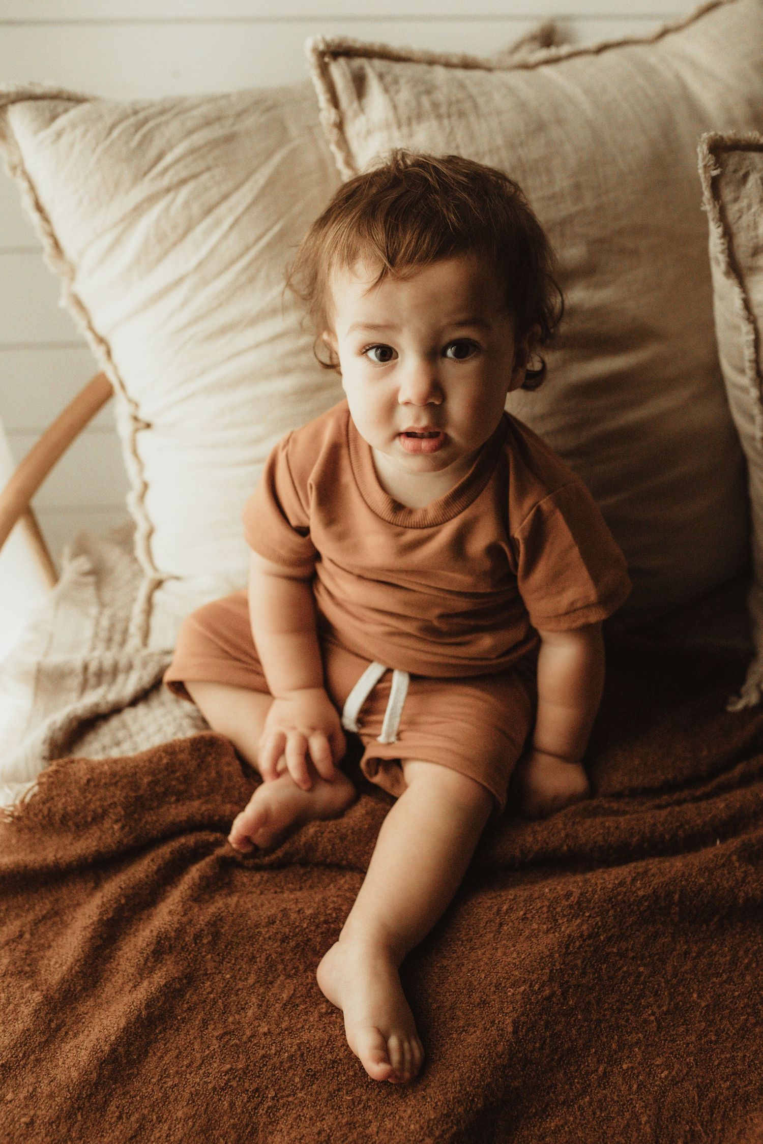Multiple shots of a young child in earth-toned clothing lounging on a brown textured blanket with beige pillows.