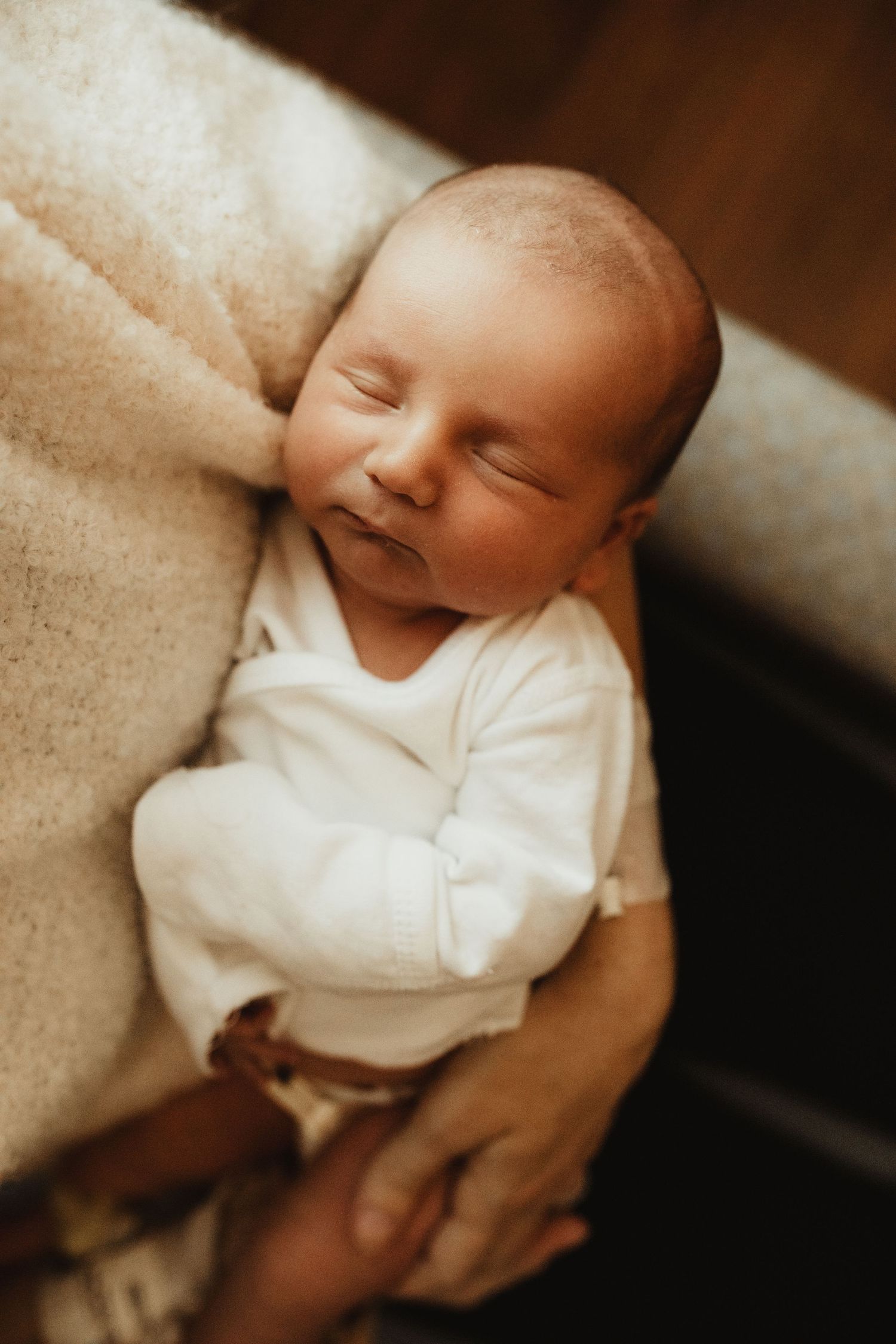 A newborn baby in white sleepwear rests peacefully on a cozy beige blanket in warm sepia lighting.