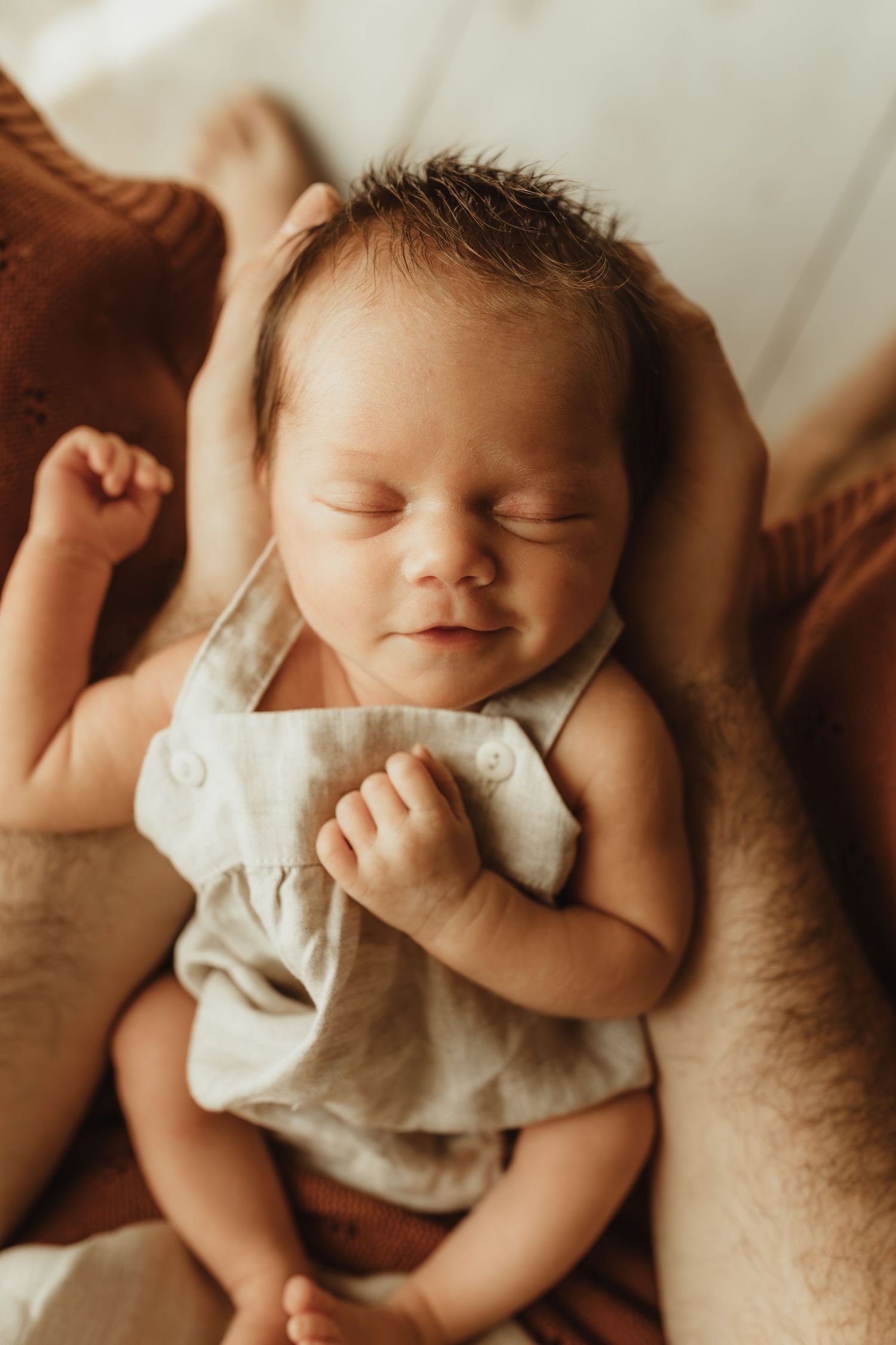 Peaceful newborn baby sleeping in a light gray linen romper, cradled in loving hands on a warm neutral background.