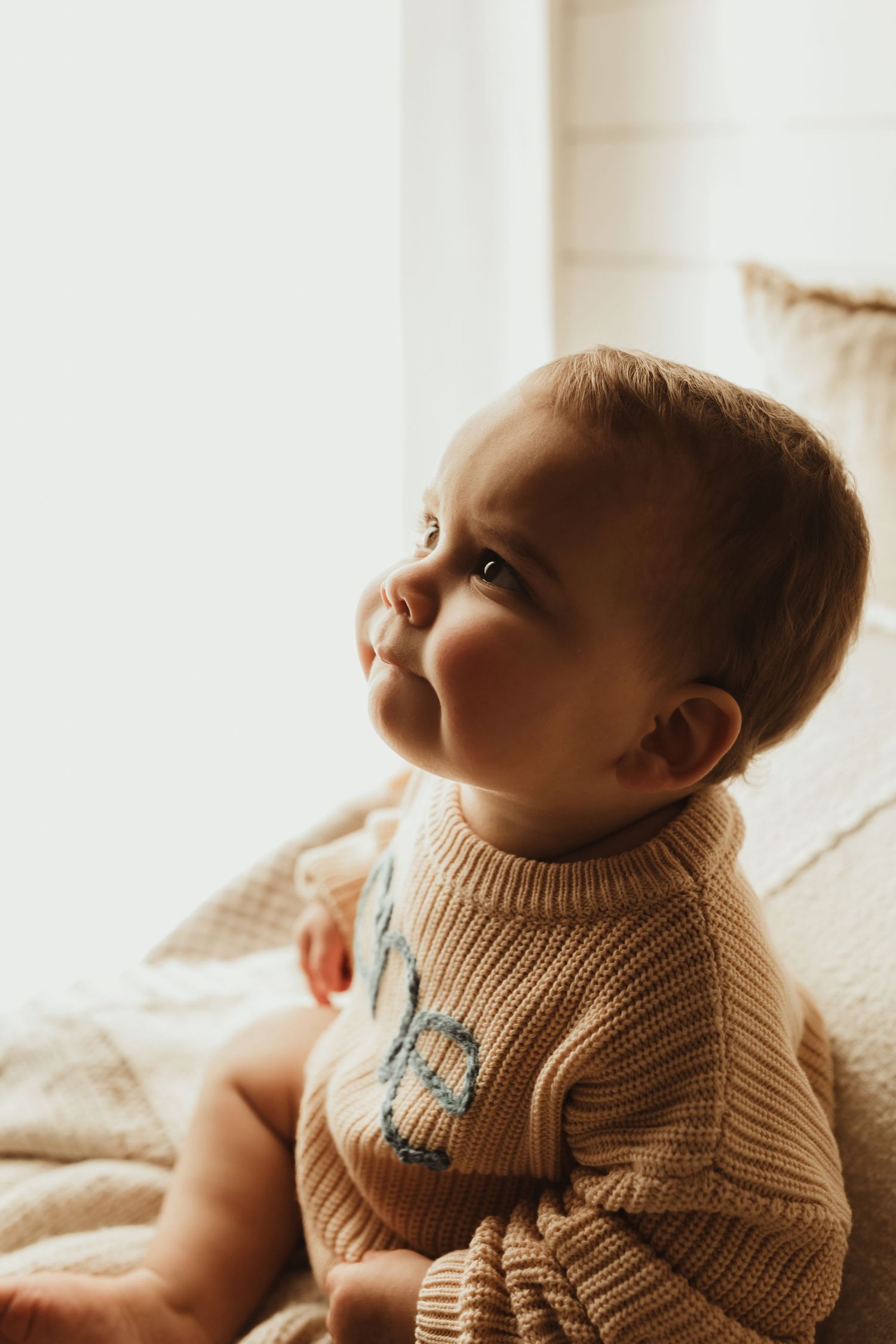 Baby in beige knit sweater lying on soft white bedding while looking up with gentle expression.