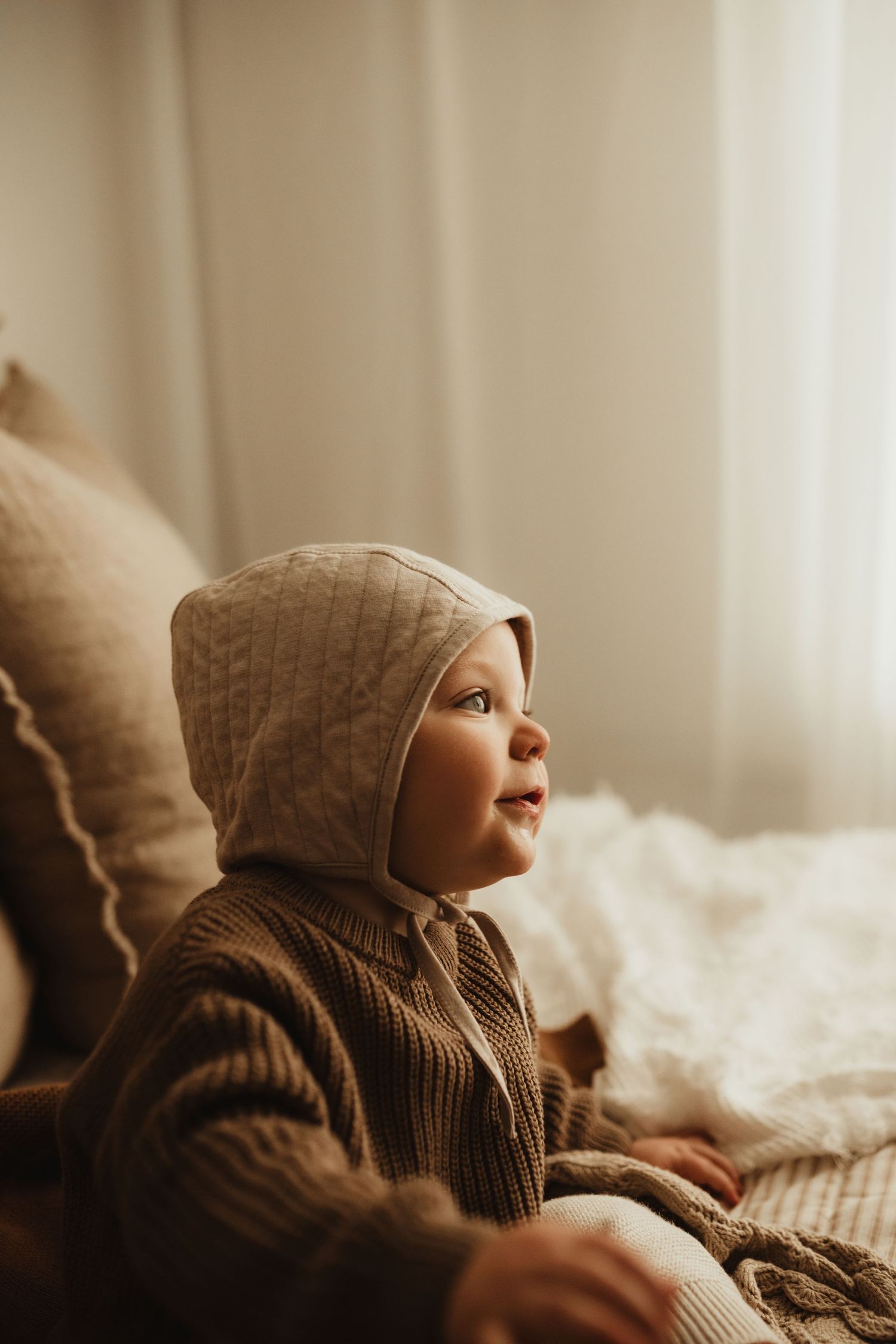 A small child in a brown knit sweater gazes upward while resting on soft cushions in warm natural lighting.