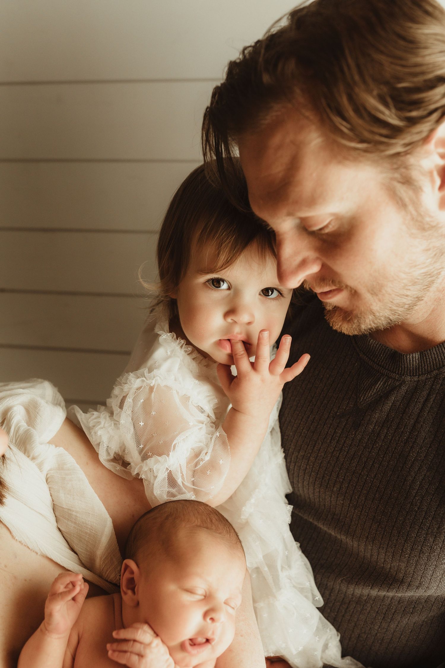Father tenderly holds toddler daughter and sleeping newborn baby during a warm family portrait session.