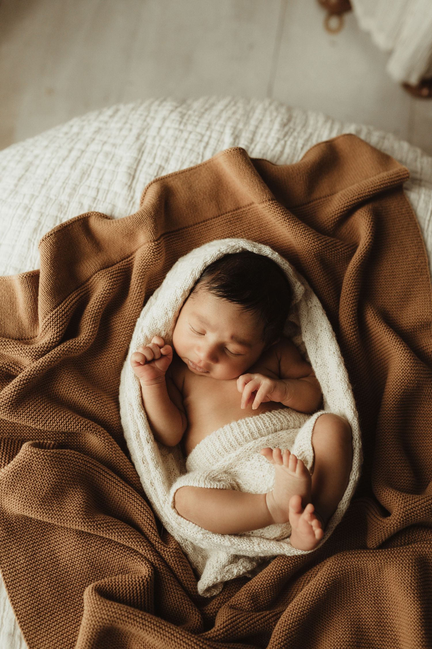 A sleeping newborn wrapped in a white knit blanket lies on a cozy brown textured blanket during a photography session.