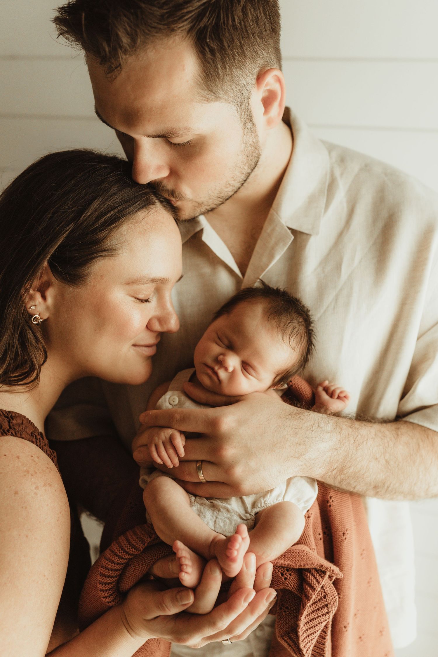 New parents tenderly hold their sleeping newborn baby, father kissing mother's head in a warm, intimate portrait.