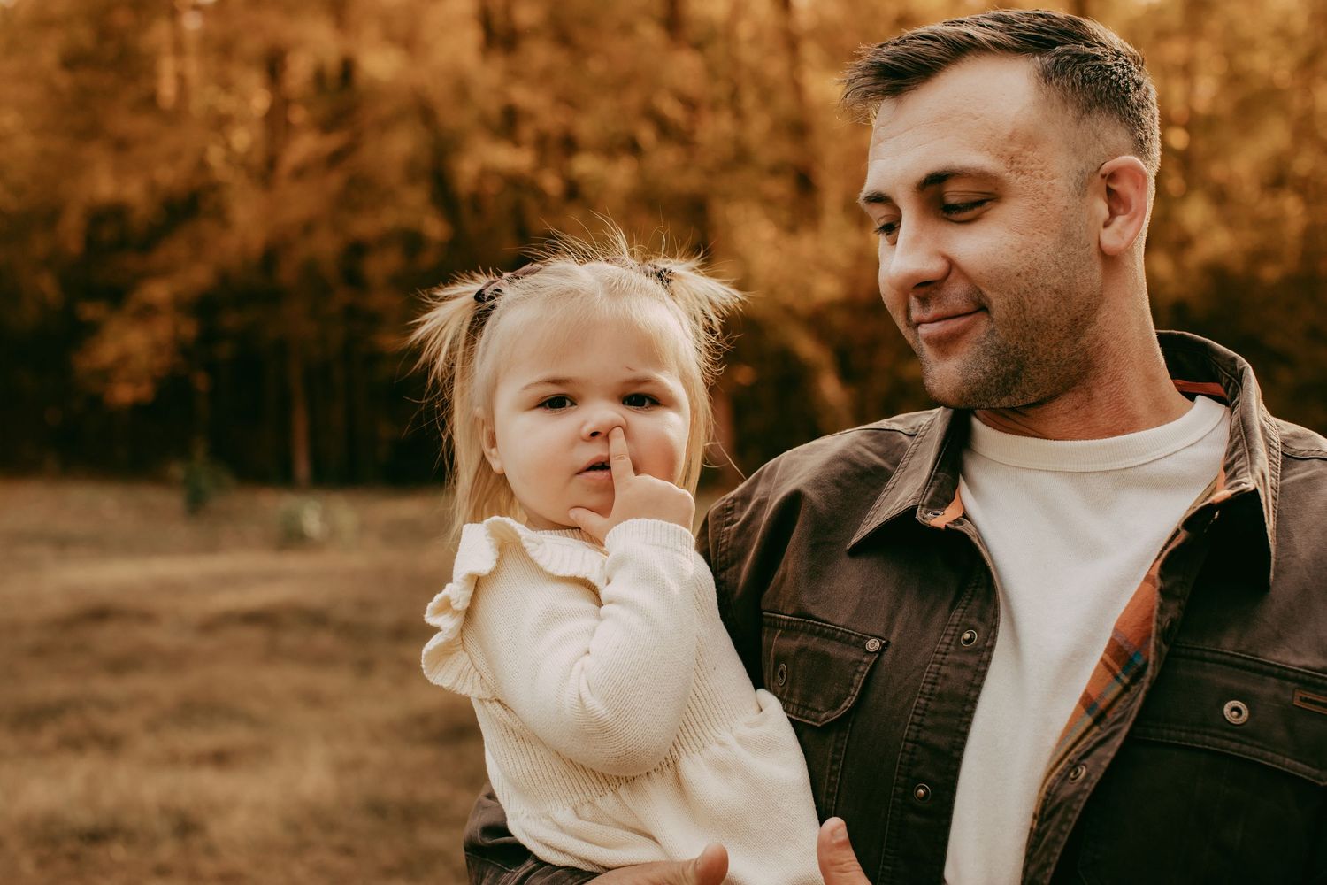 A father holds his toddler daughter outdoors in an autumn forest setting, both dressed in warm casual clothing.