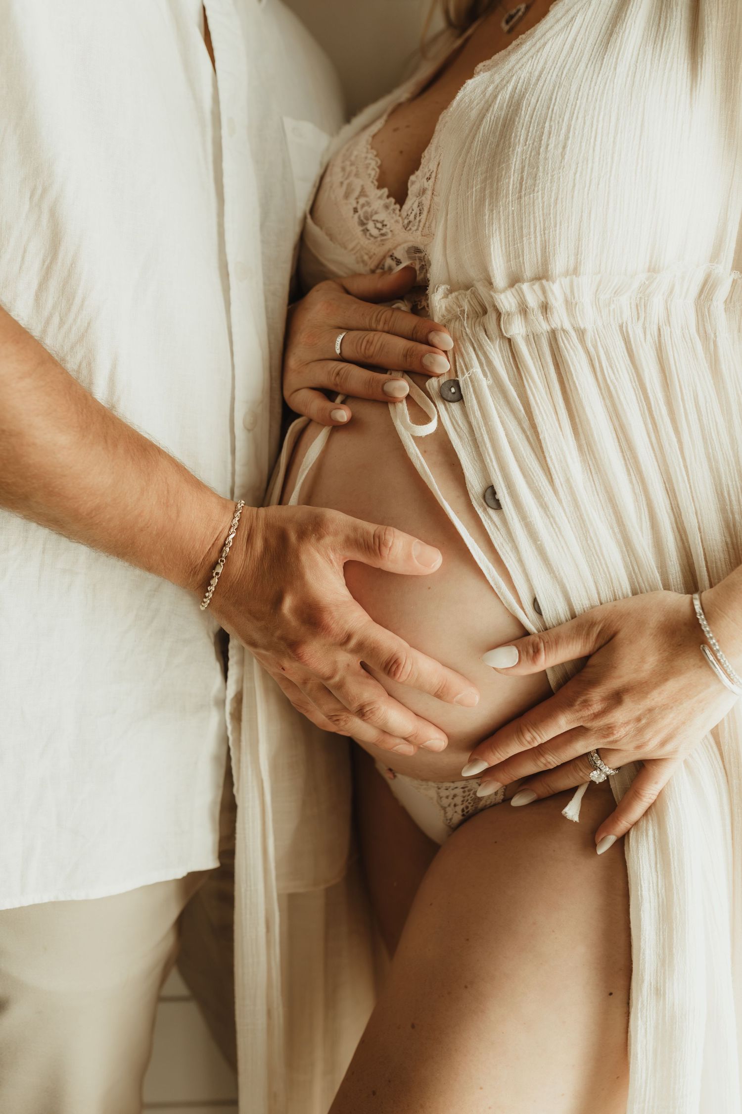 Couple tenderly cradling pregnant belly, wearing neutral beige tones, with rings and bracelet visible.