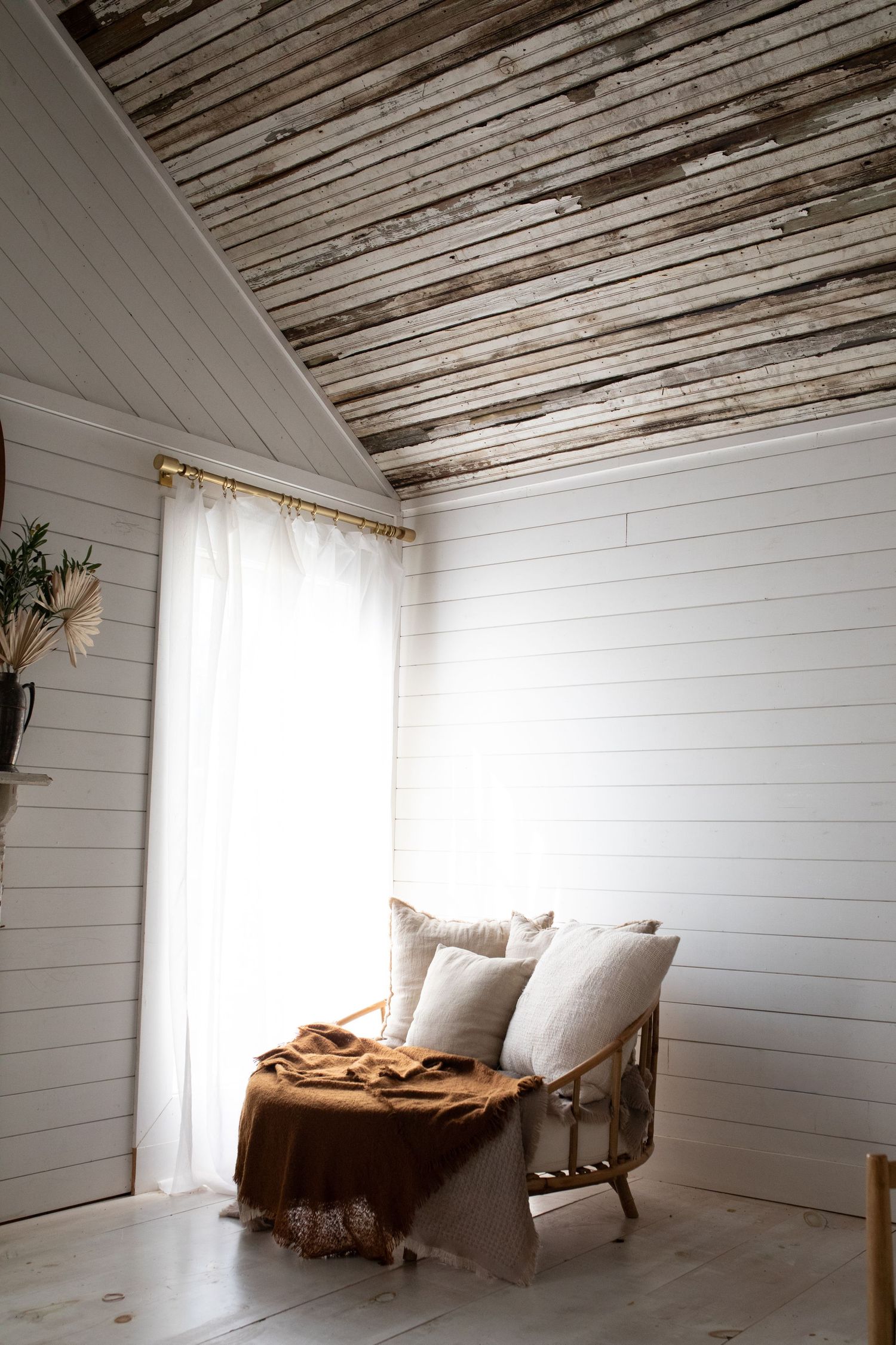 White shiplap walls and rustic wooden ceiling create a minimalist bedroom space with natural light streaming through windows.