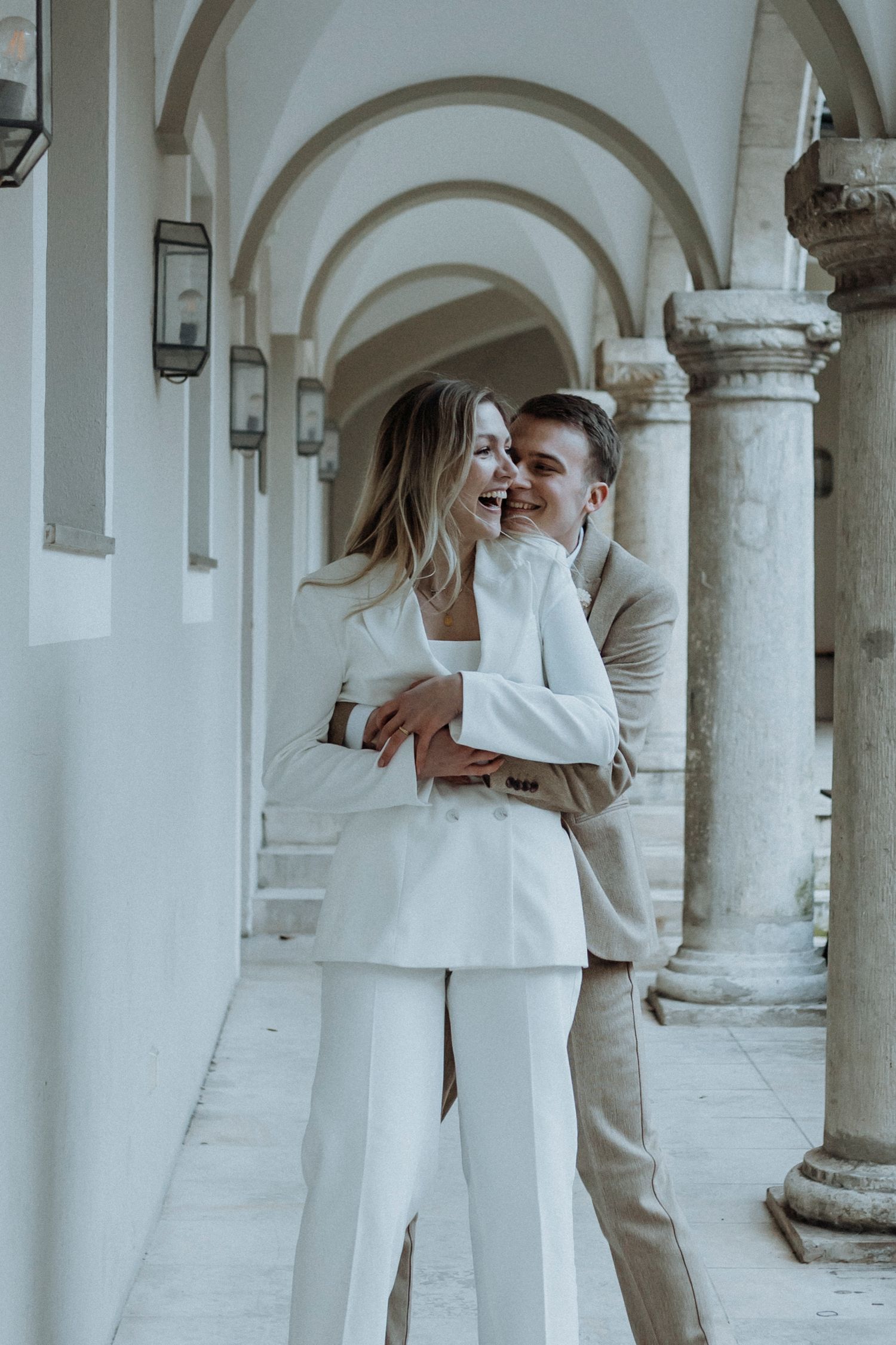 Romantic couple embracing in an elegant arched colonnade wearing matching light colored outfits.