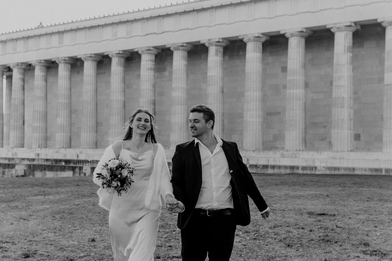 Couple walking hand in hand beside a grand neoclassical building with columns.