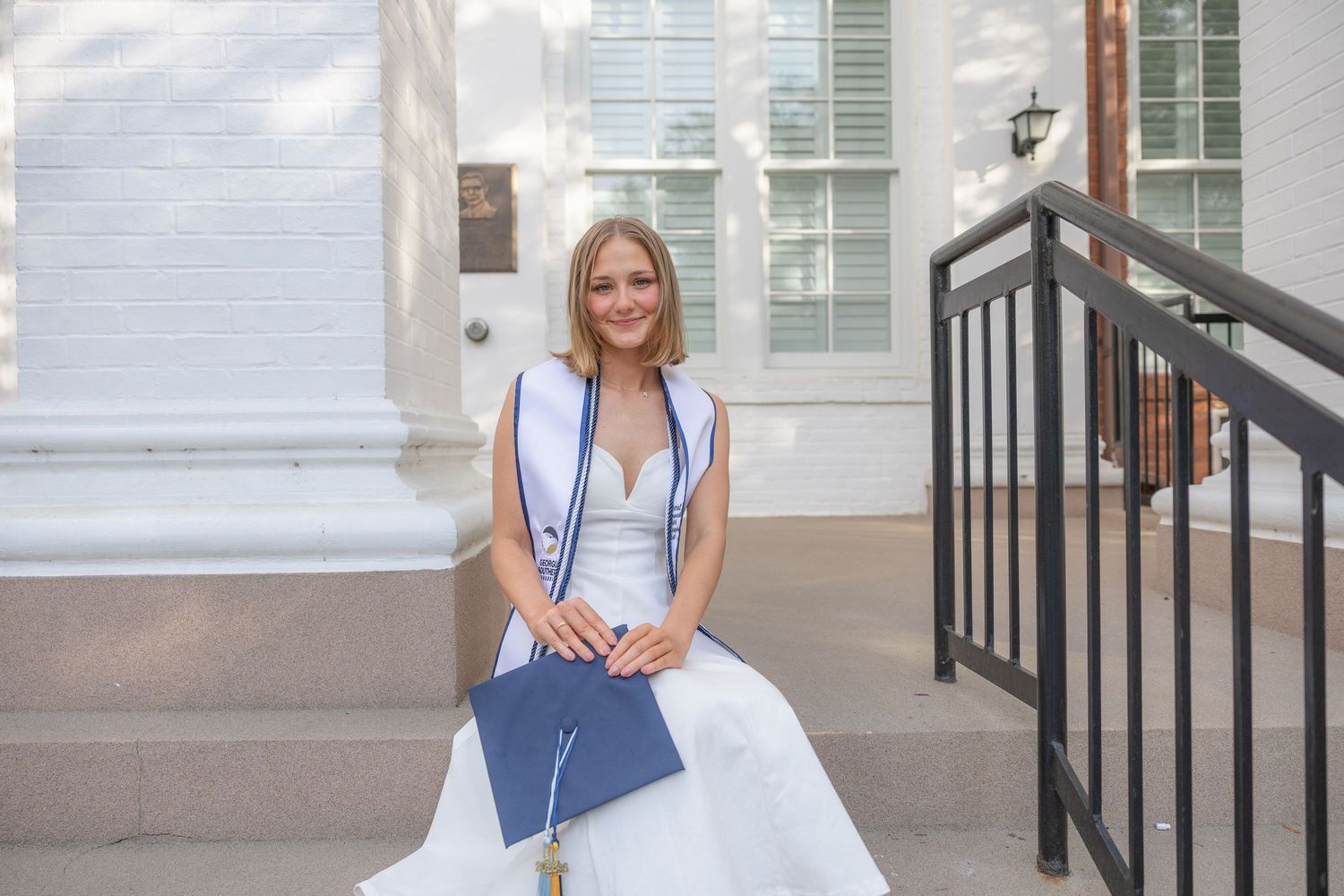 Smiling graduate in white gown holding diploma sitting on steps outside a historic building.