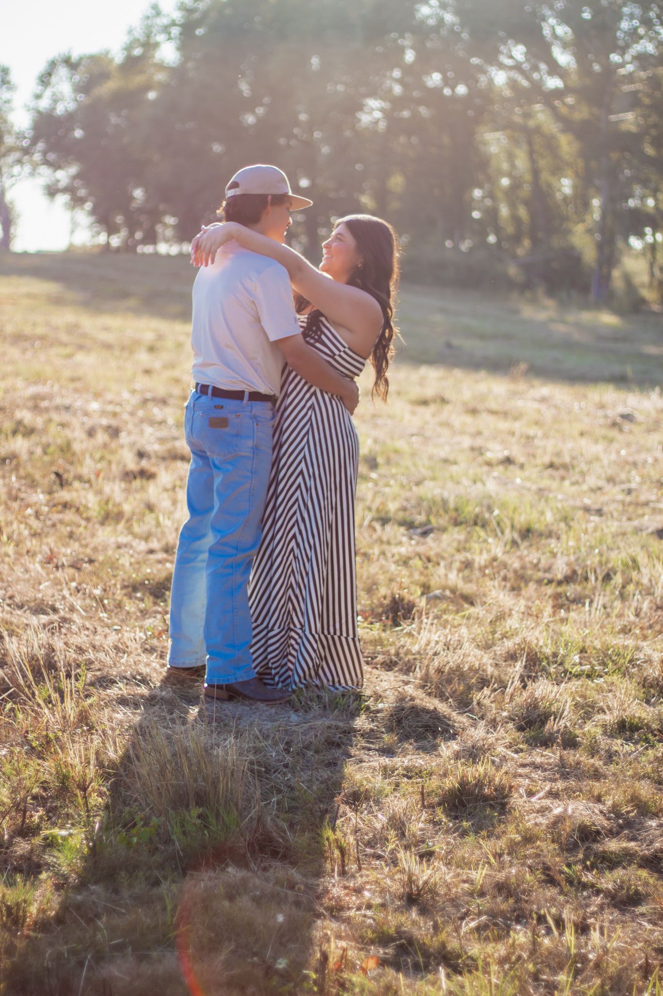 Couple embracing in golden sunlit field, woman in striped maxi dress, romantic outdoor portrait.
