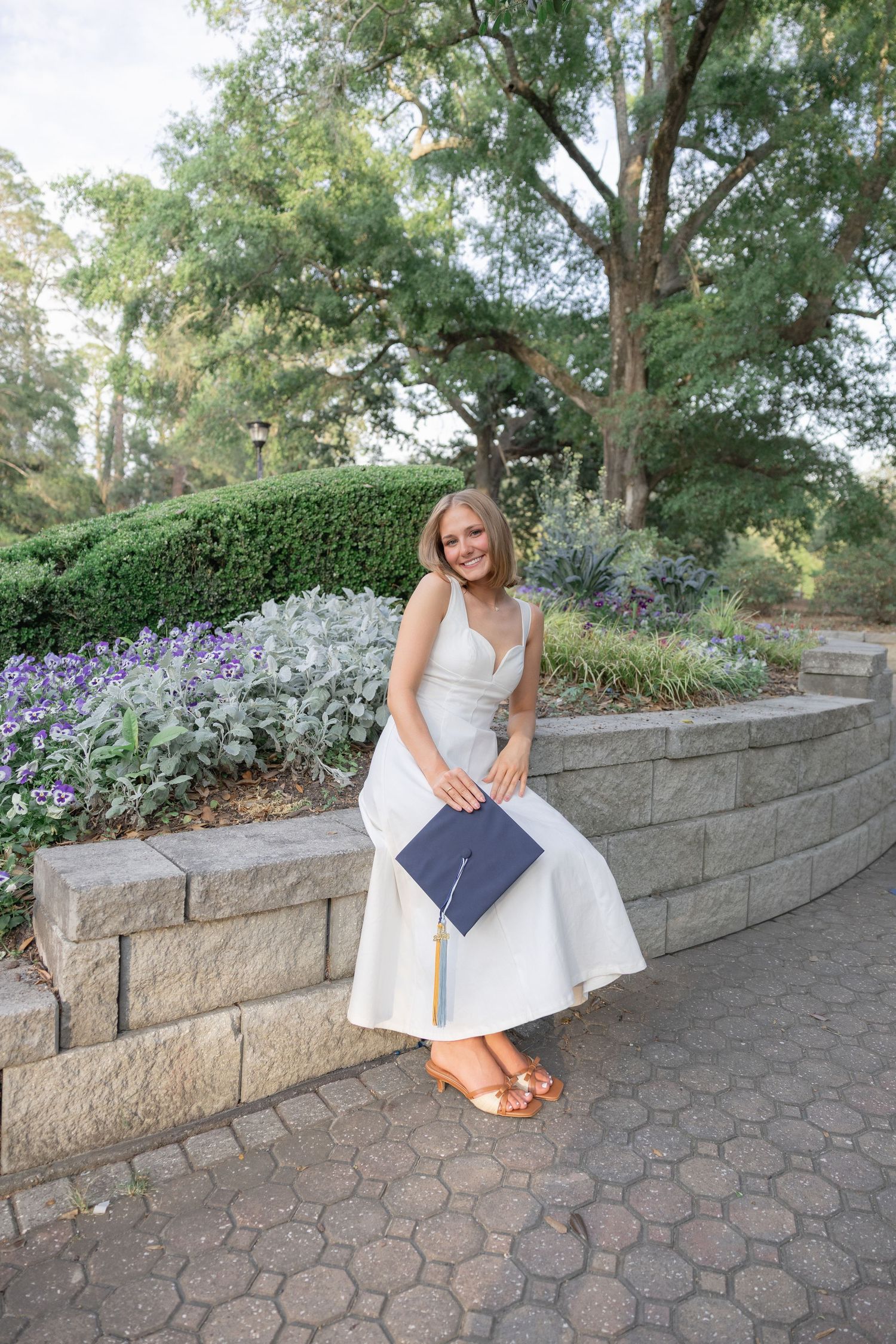Smiling graduate in white dress holding navy cap poses by stone wall in lush garden setting.