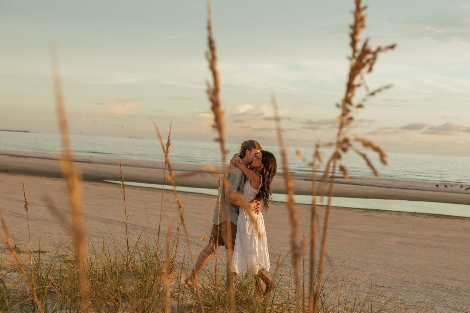 Romantic couple embracing on sandy beach at golden sunset with tall sea oats in foreground.
