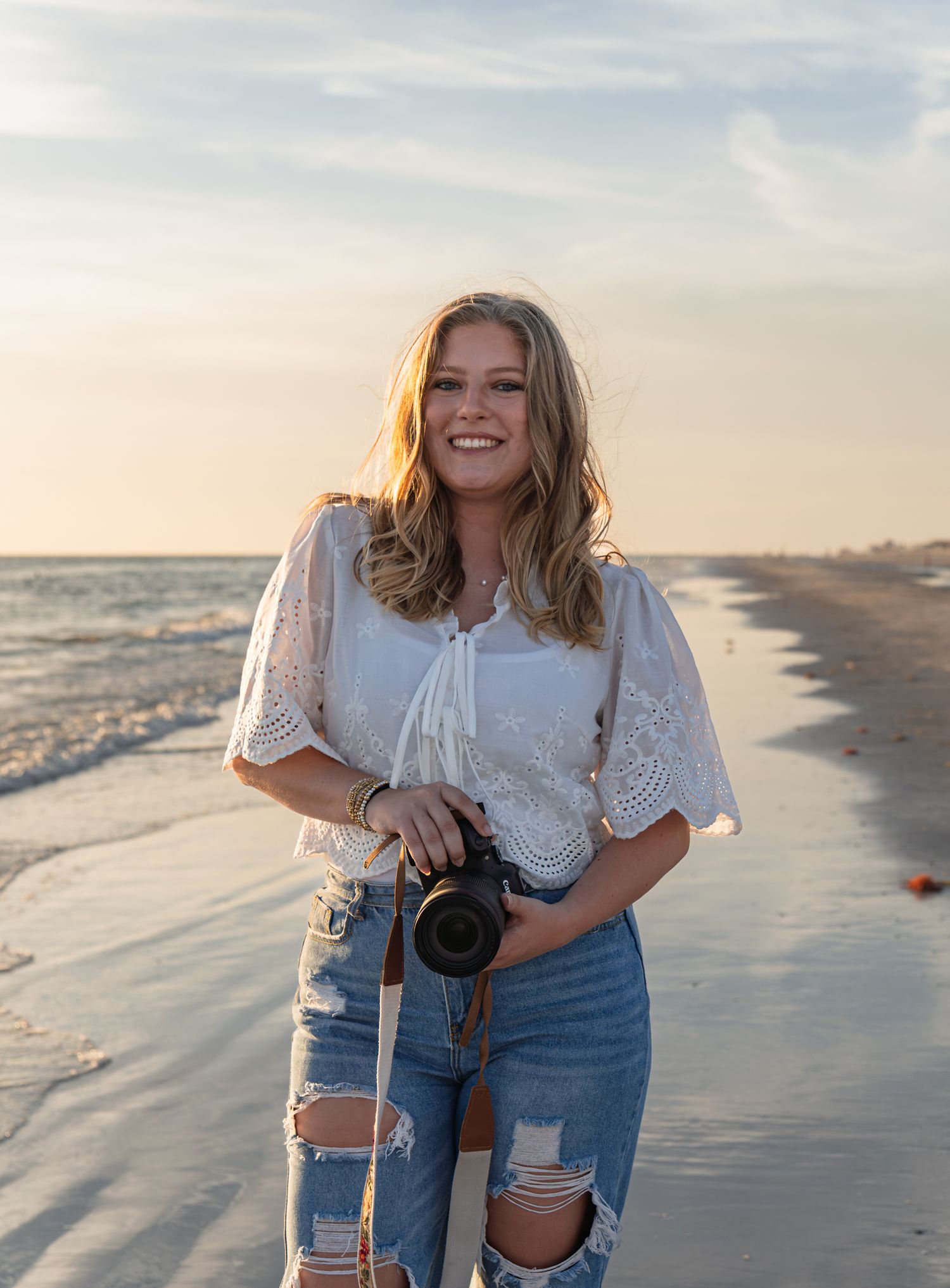 Smiling female photographer holding a camera on a sunny beach in ripped jeans and white top.
