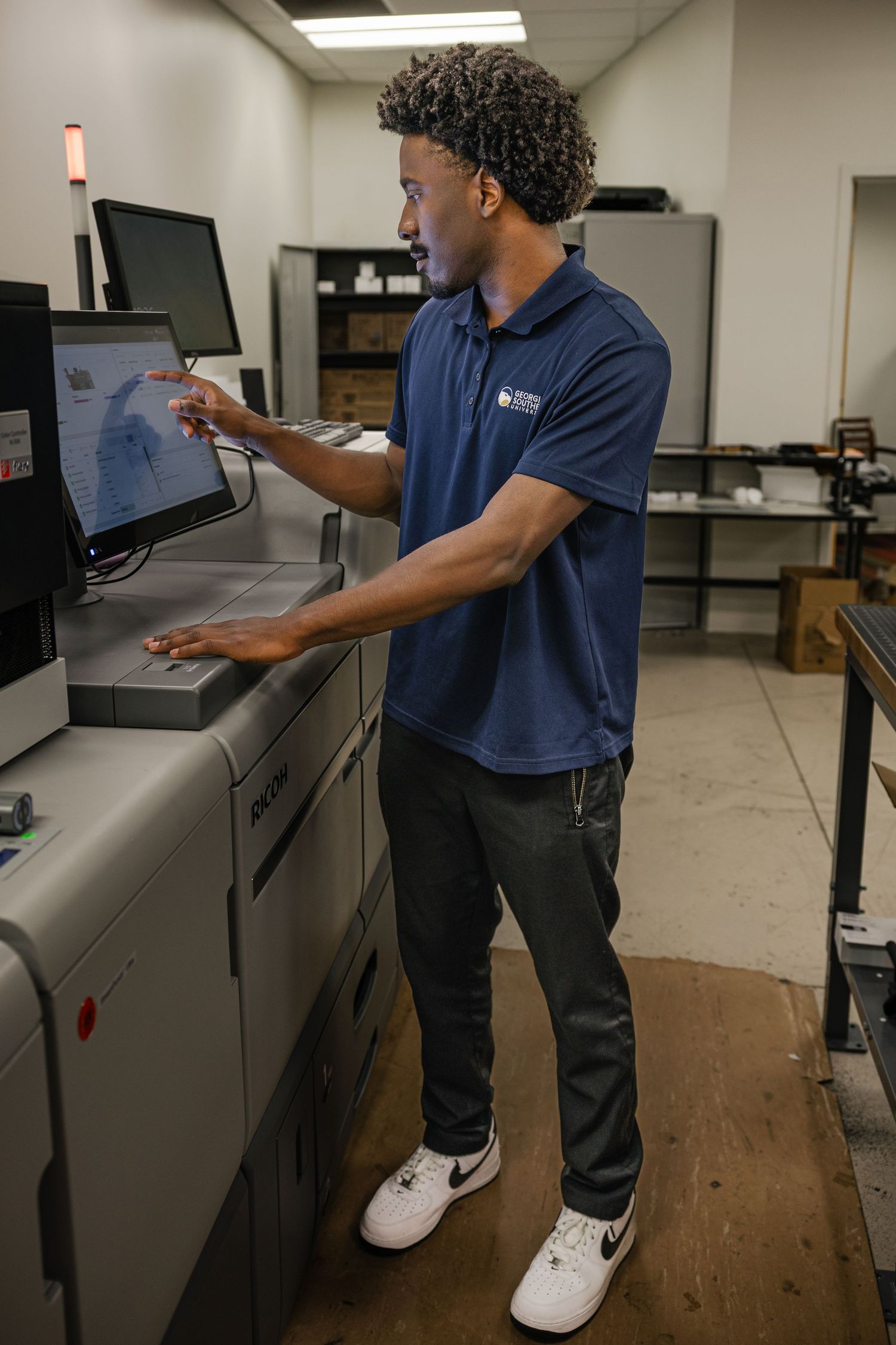 A young man in a blue polo shirt operates a large digital printing machine in a professional print shop.
