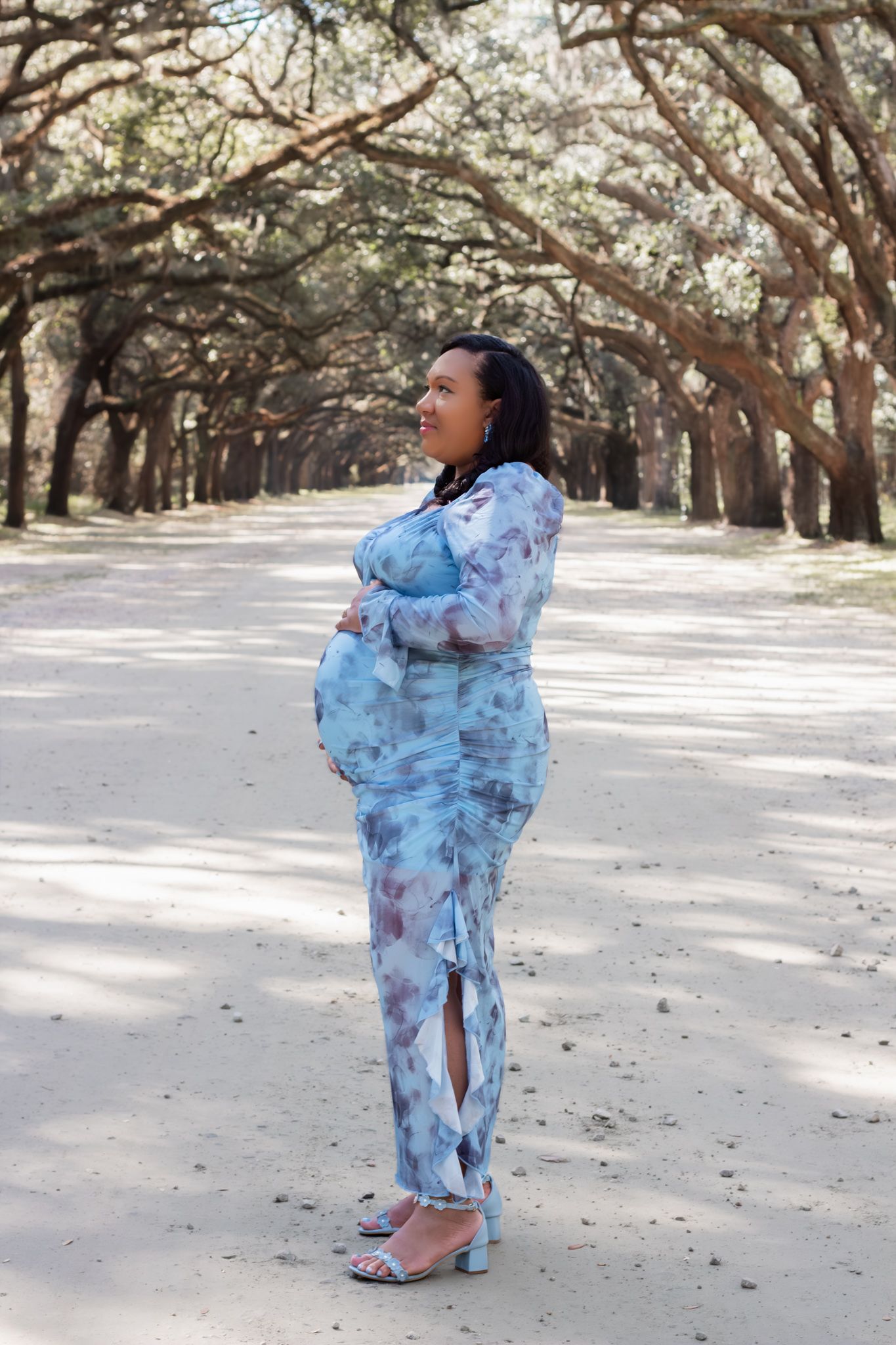 Pregnant woman in floral blue gown stands on tree-lined sandy path in serene park.