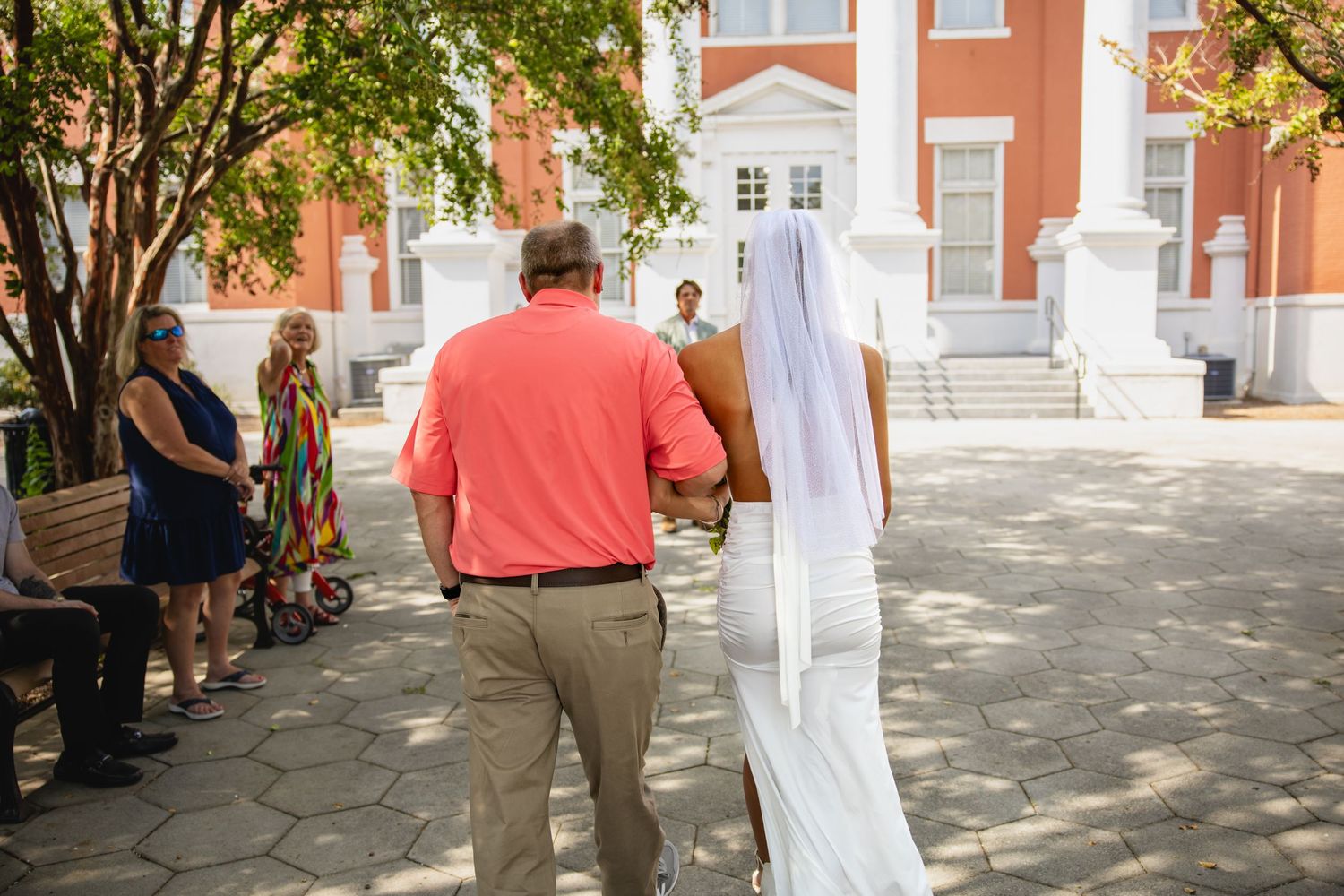 Bride in white gown and veil walks with man in coral shirt toward red brick building.