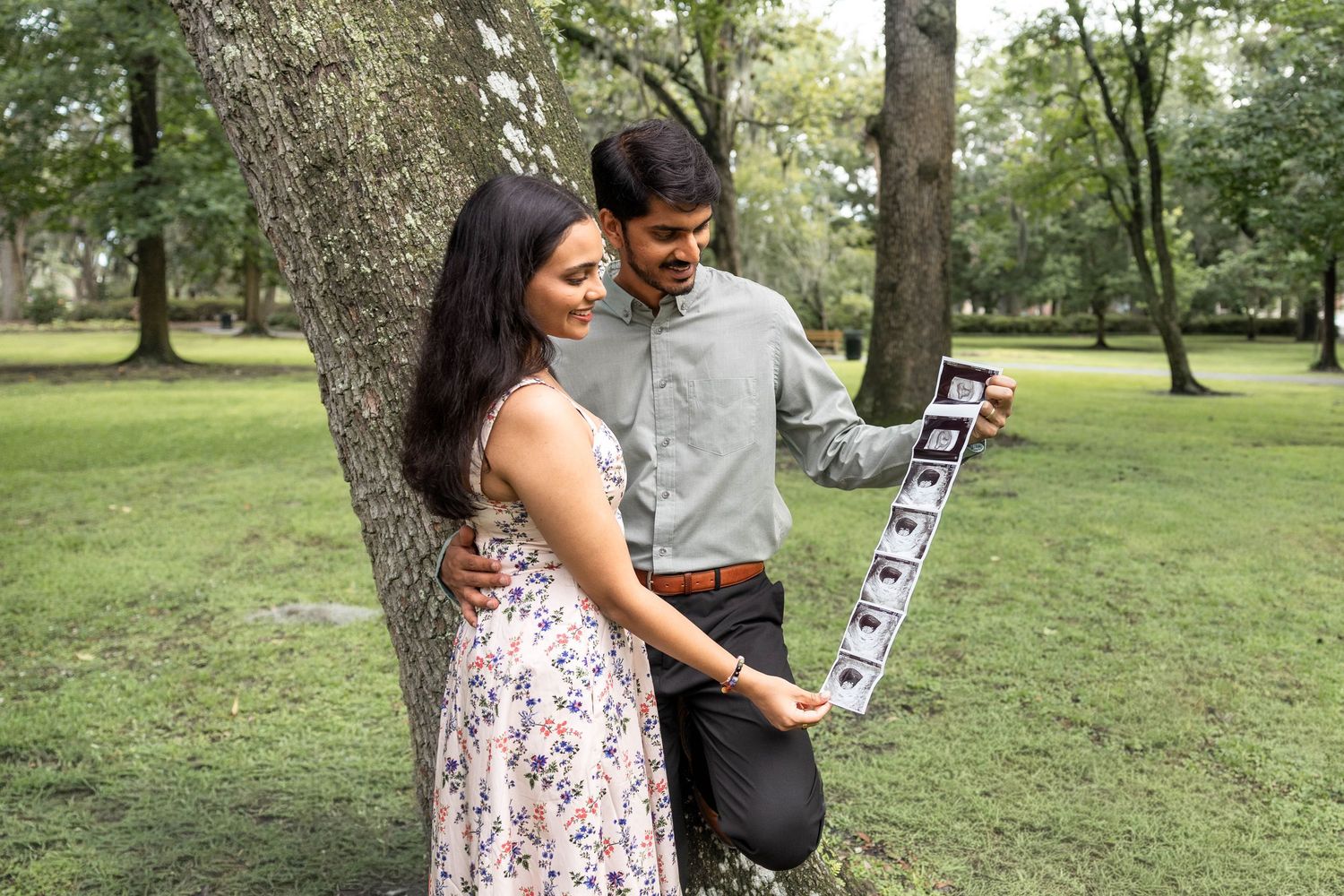 Couple leaning against tree in park looking at ultrasound photos together.