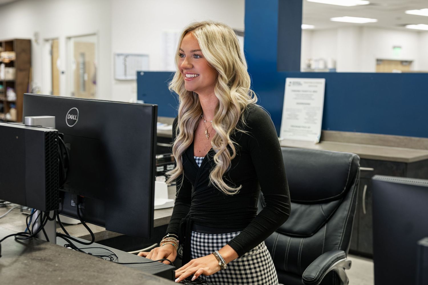 A smiling blonde woman works at a Dell computer workstation in a modern office environment.