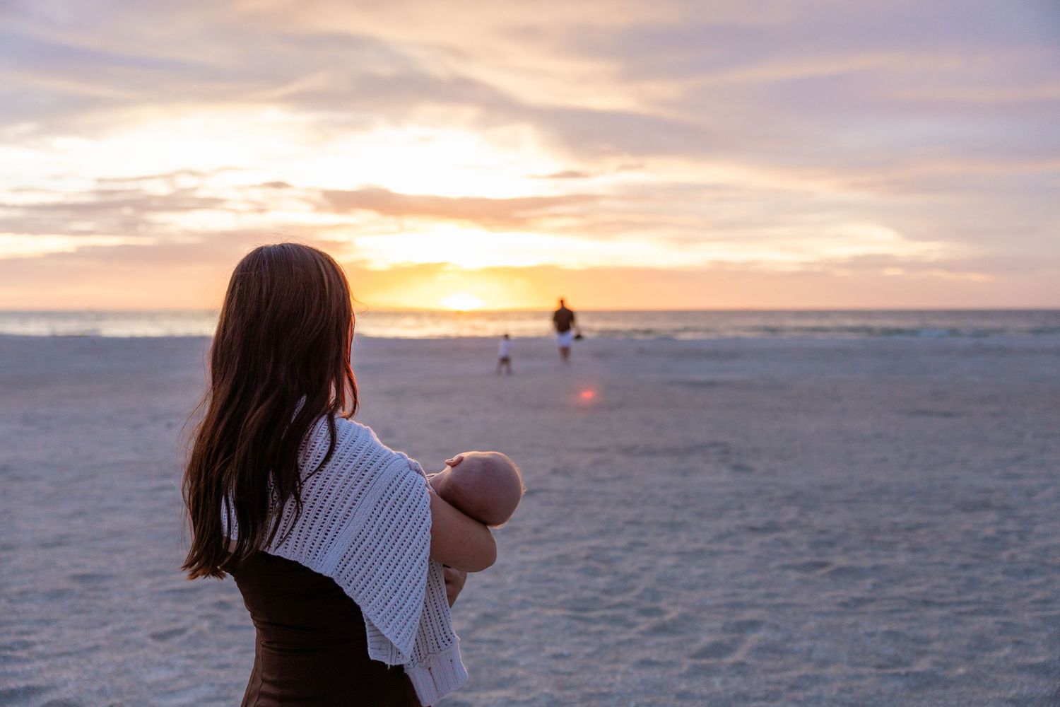 Mother holding newborn baby watches family on beach during stunning golden hour sunset.