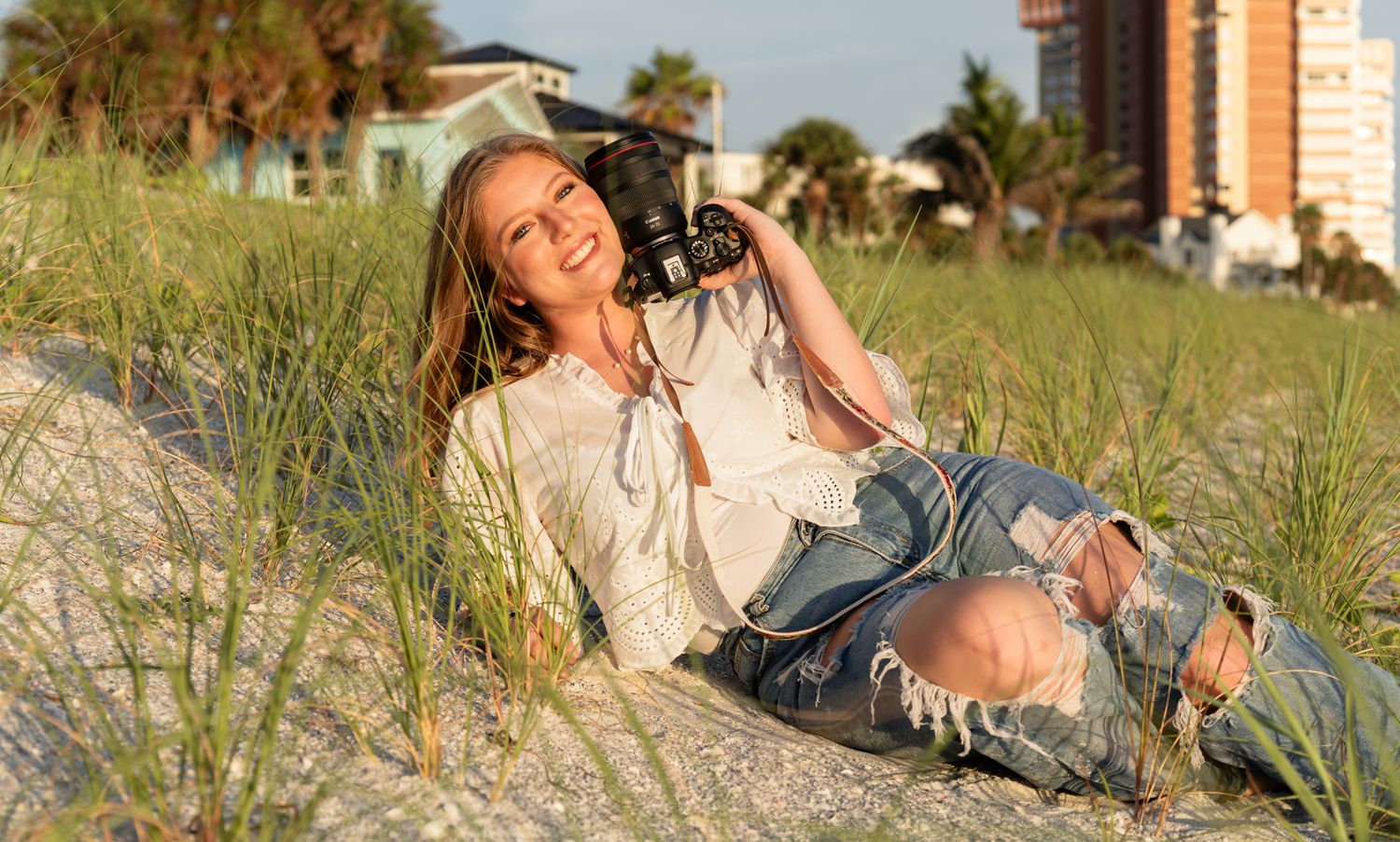 Young woman with camera relaxing on sandy beach dunes surrounded by green grass at golden hour.