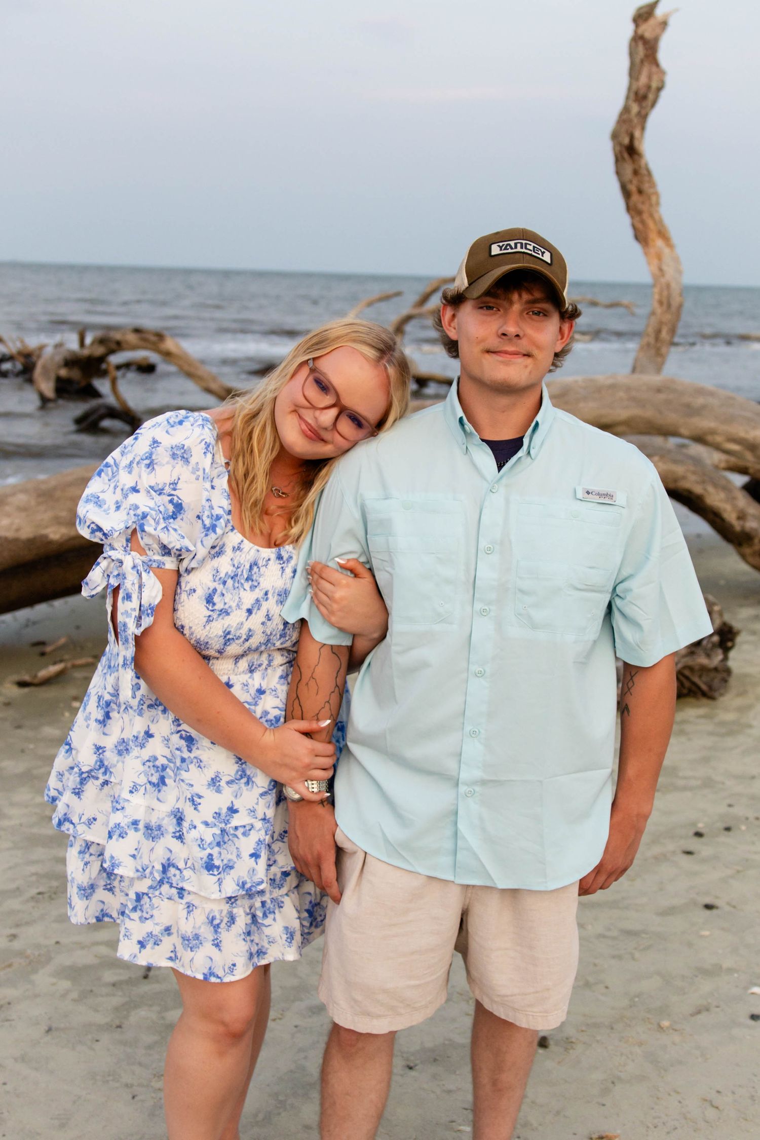 A young couple poses together on a beach with driftwood and ocean waves in the background.