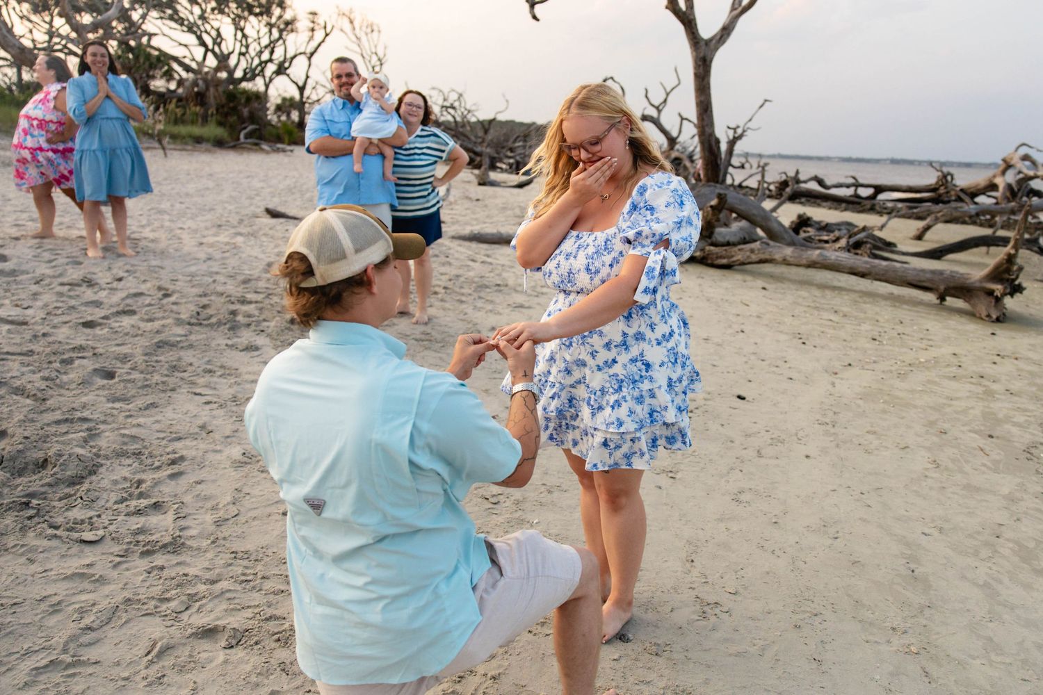 Emotional beach proposal with man on one knee as crowd of family watches in background.