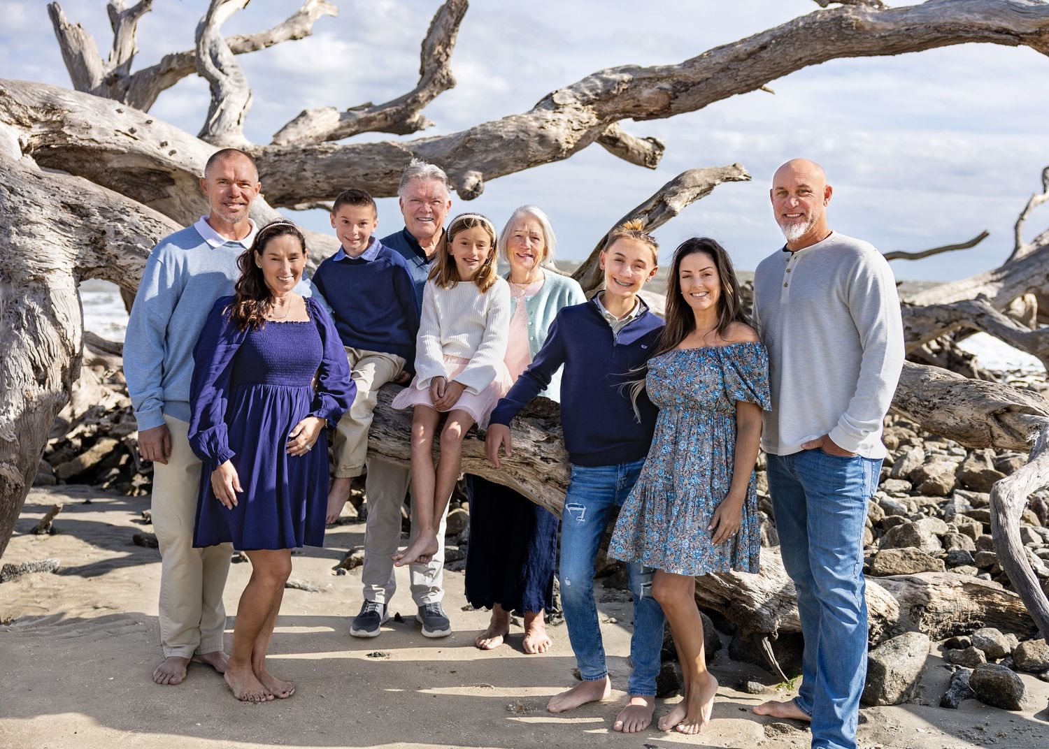 A large family group of eight people poses together among driftwood on a scenic beach.