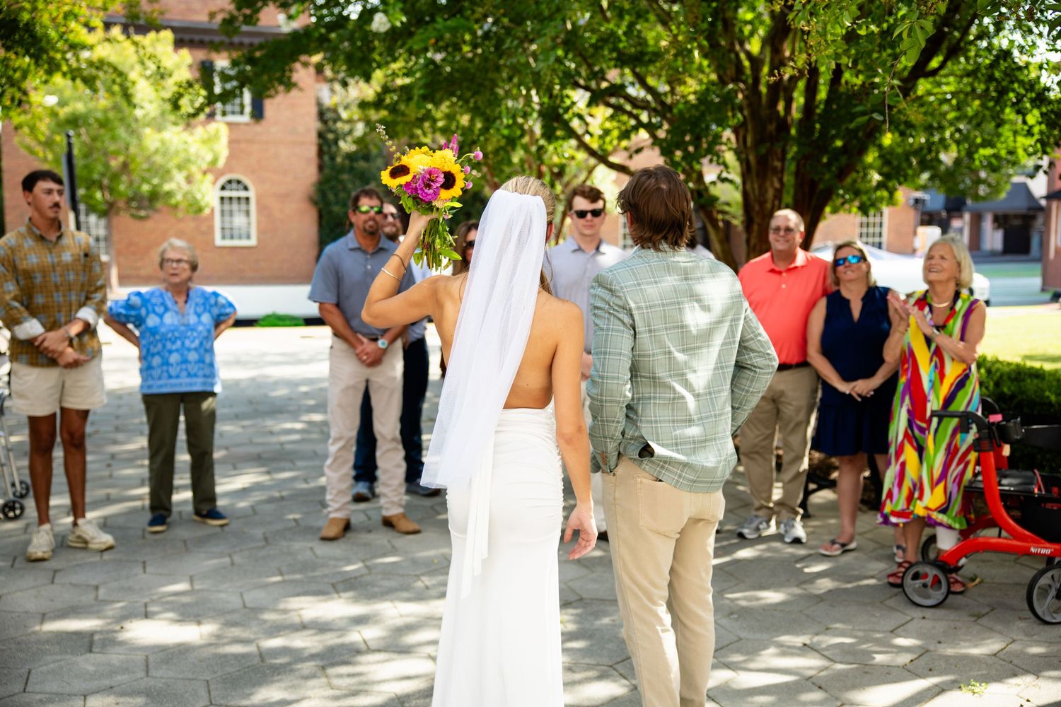 Bride holding colorful bouquet approaches smiling groom surrounded by wedding guests in outdoor plaza.