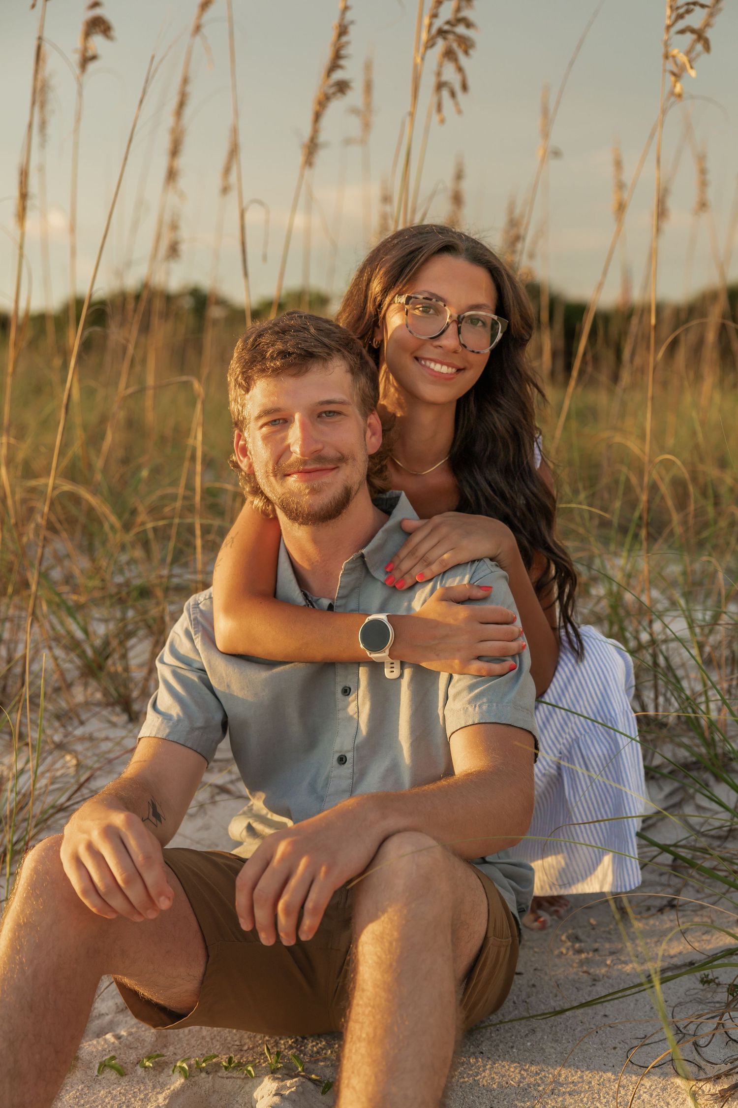 Smiling couple sitting together in beach sand dunes at sunset, woman hugging man from behind.