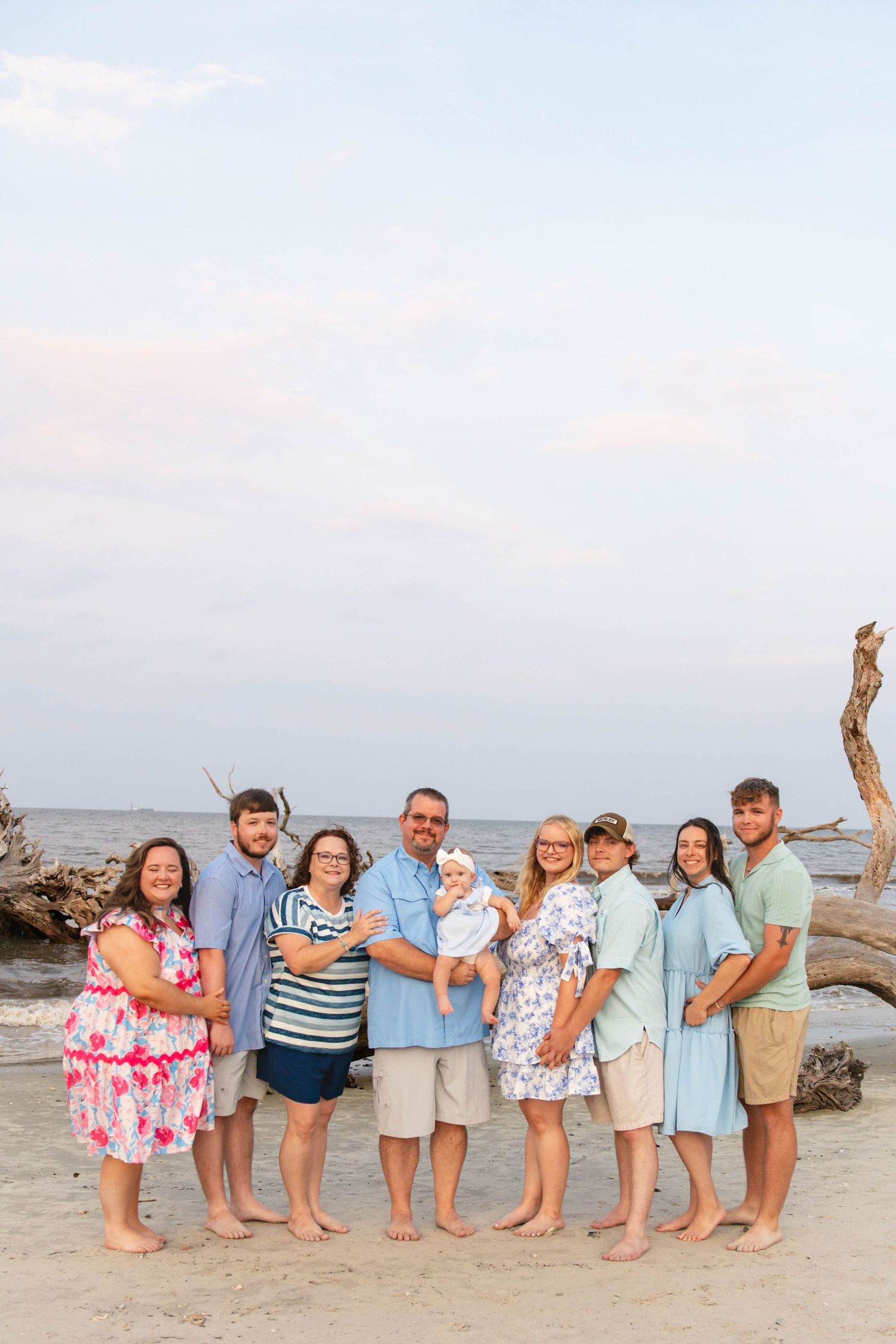 Extended family of eight poses barefoot on beach at sunset with driftwood behind them.