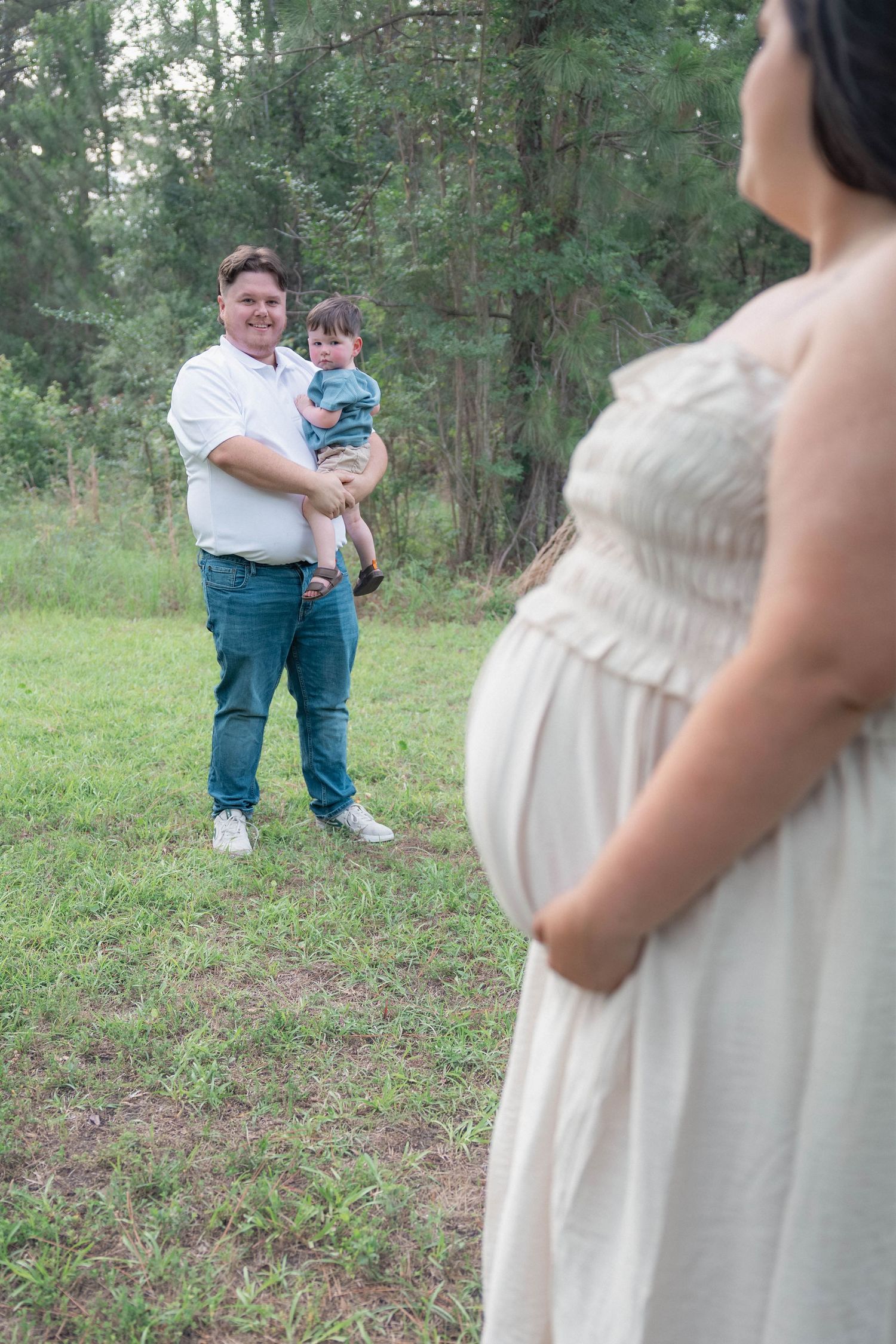 Father holding toddler smiling at pregnant mother in a lush green outdoor setting.