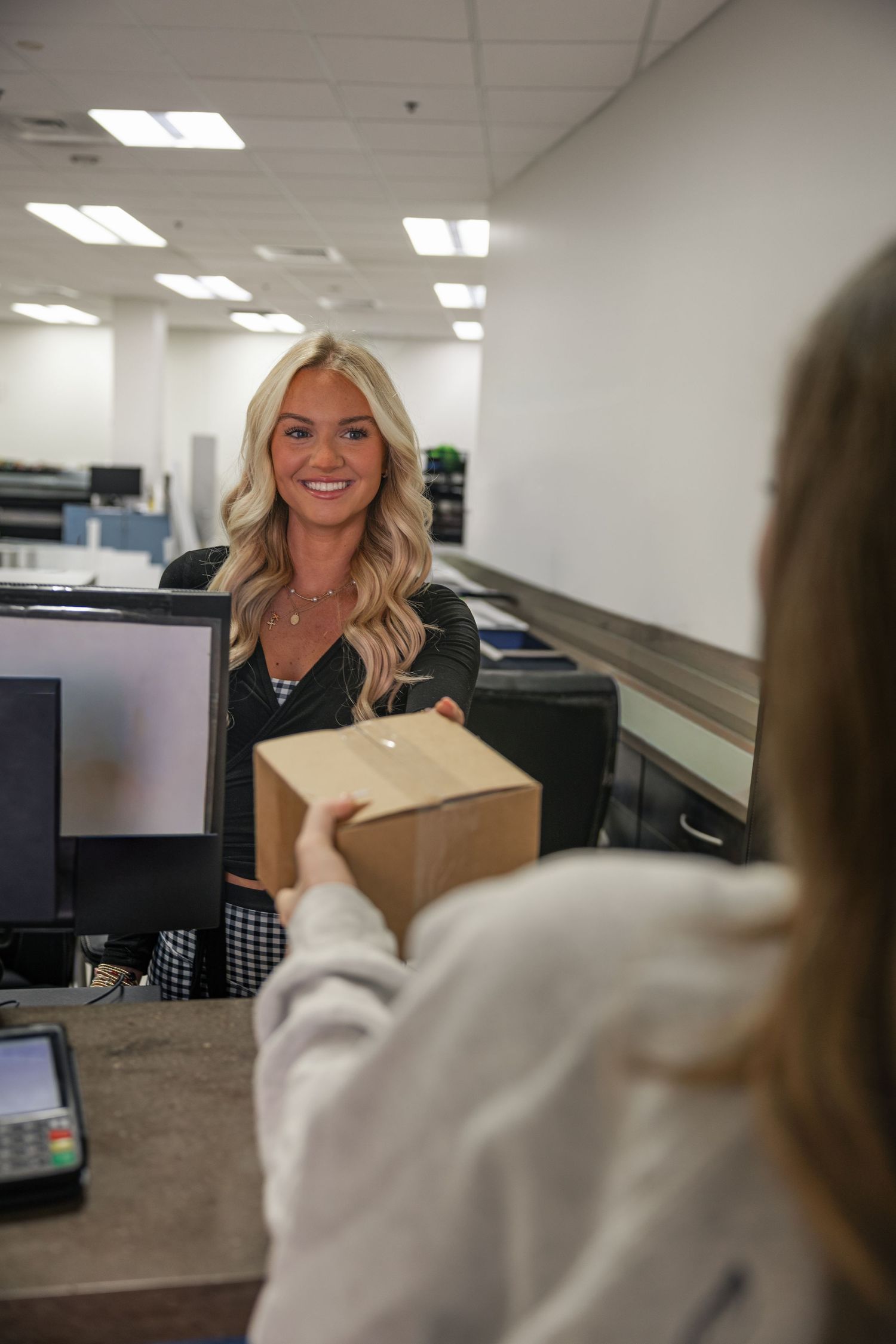 A cheerful blonde female clerk receives a brown package from a customer at a campus mail center.